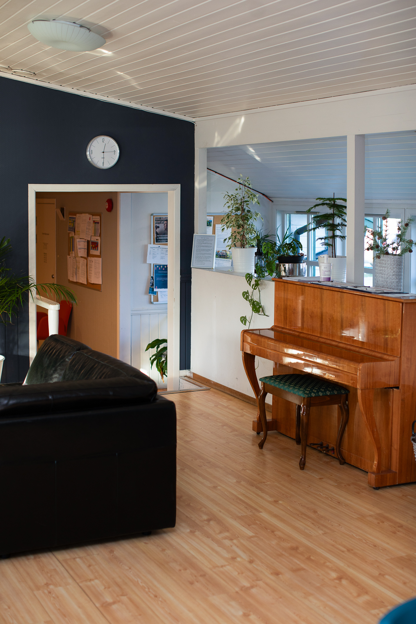 Foto: Et piano står inntil en halvvegg. Over kan man se inn i neste rom. Her er det åpent og luftig med grønne planter og sofa.