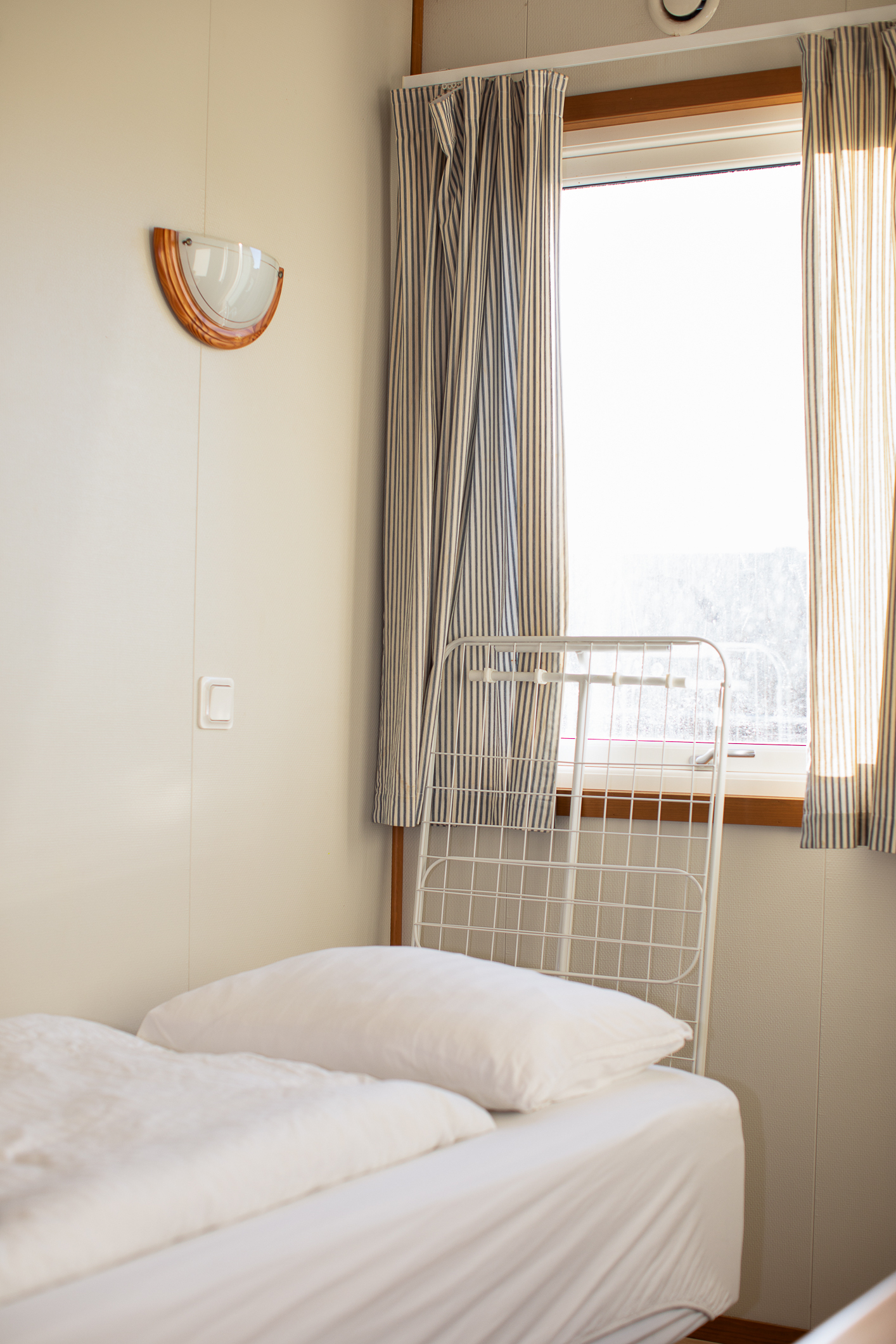 Foto: Enkel seng med hvitt sengetøy i et lyst rom. På veggen et stort vindu.