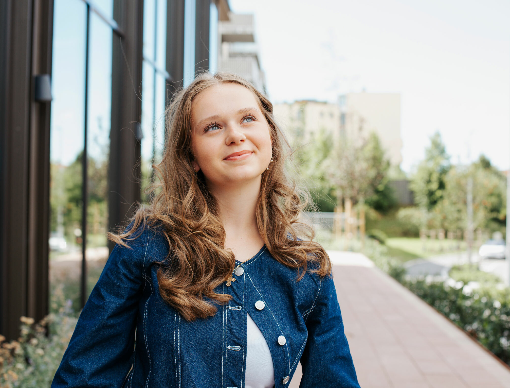 Foto: Tenåringsjente med langt, mørkeblondt hår ser opp mot himmelen. Hun har på seg en mørk blå dongerijakke og står utenfor et høyt bygg med mye glass.