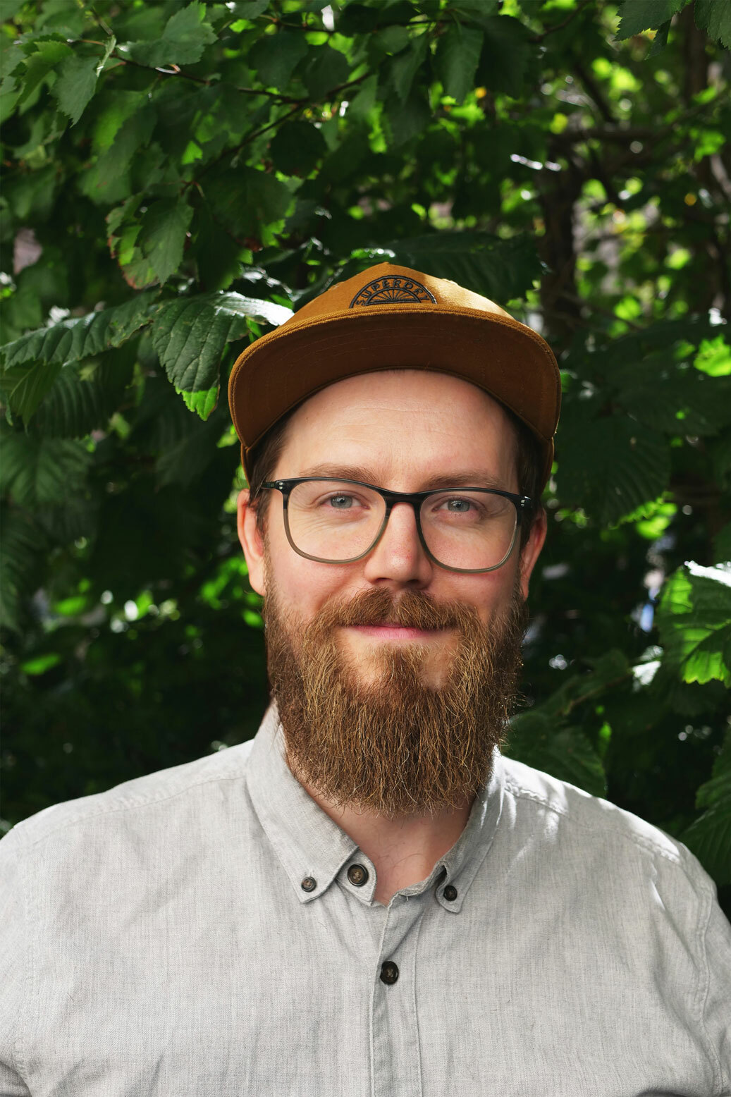 Foto: Mann sent i 20-årene med briller og skjegg. Han har på seg en brun caps og står foran en stor, grønn busk.