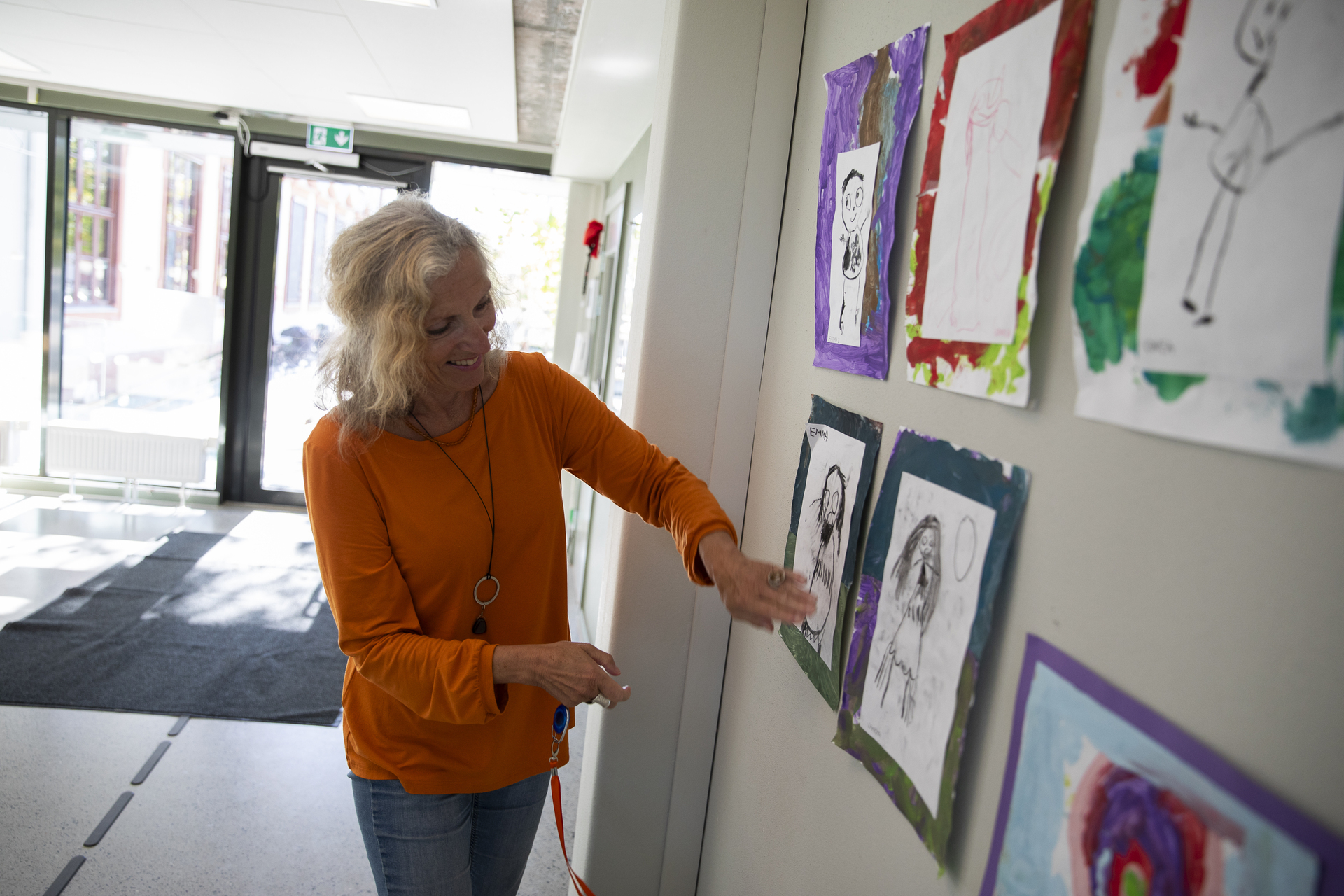 Foto: Kvinne med gråblondt, krøllete hår ser og peker på flere malerier laget av barn.