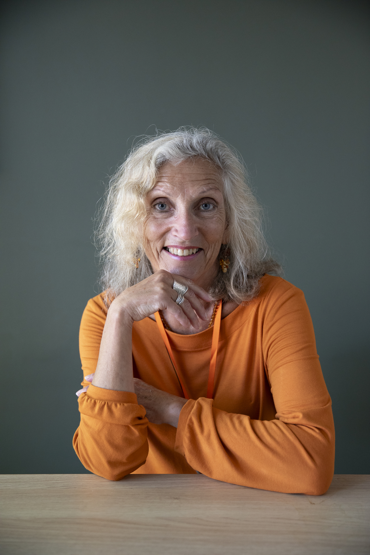 Foto: Kvinne i oransje topp og gråblondt, krøllete hår. Sitter inntil et bord og lener hodet mot ene hånden.