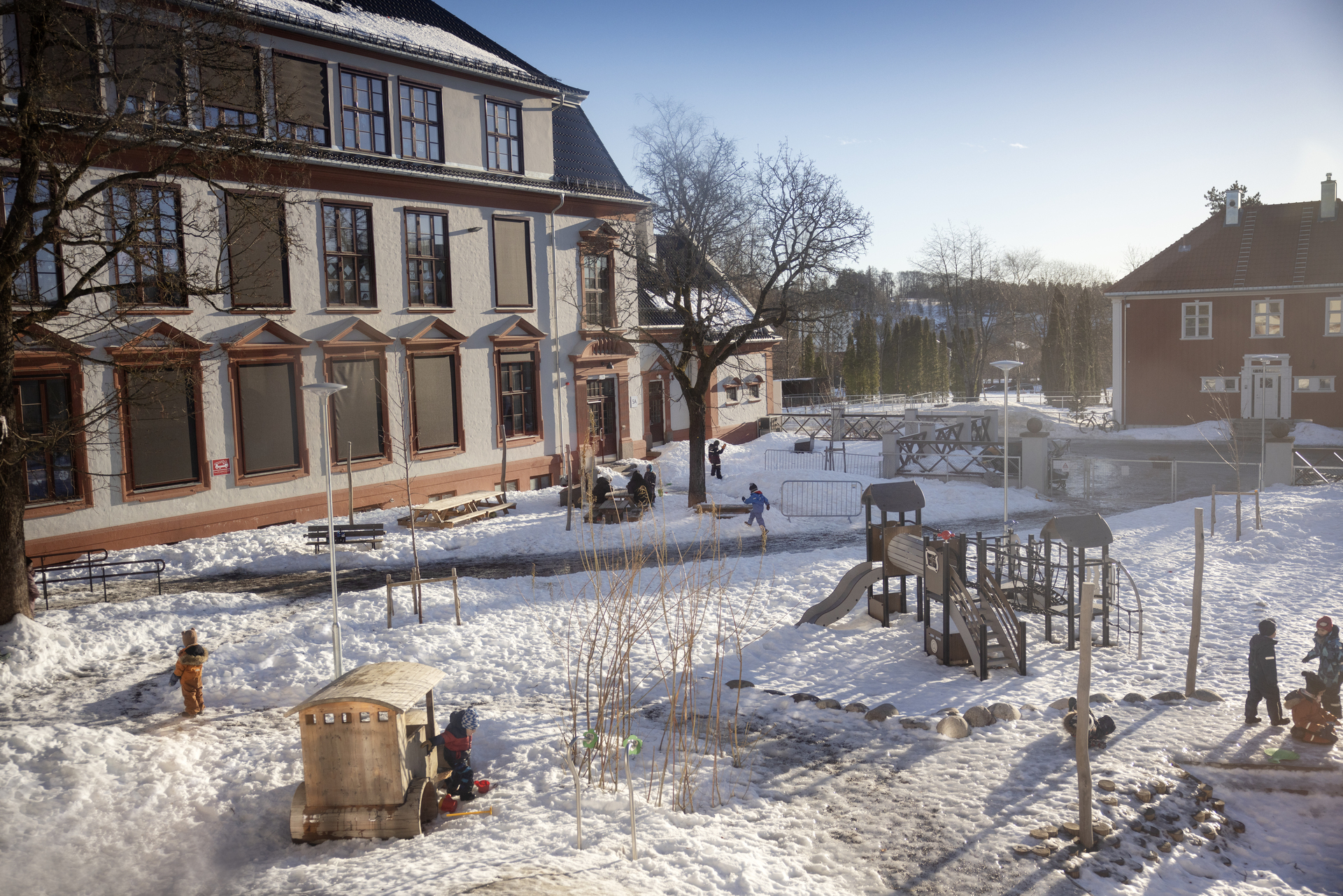 Foto: Utendørs bilde av lekeplass med mye snø foran en ærverdig bygning i fire etasjer.