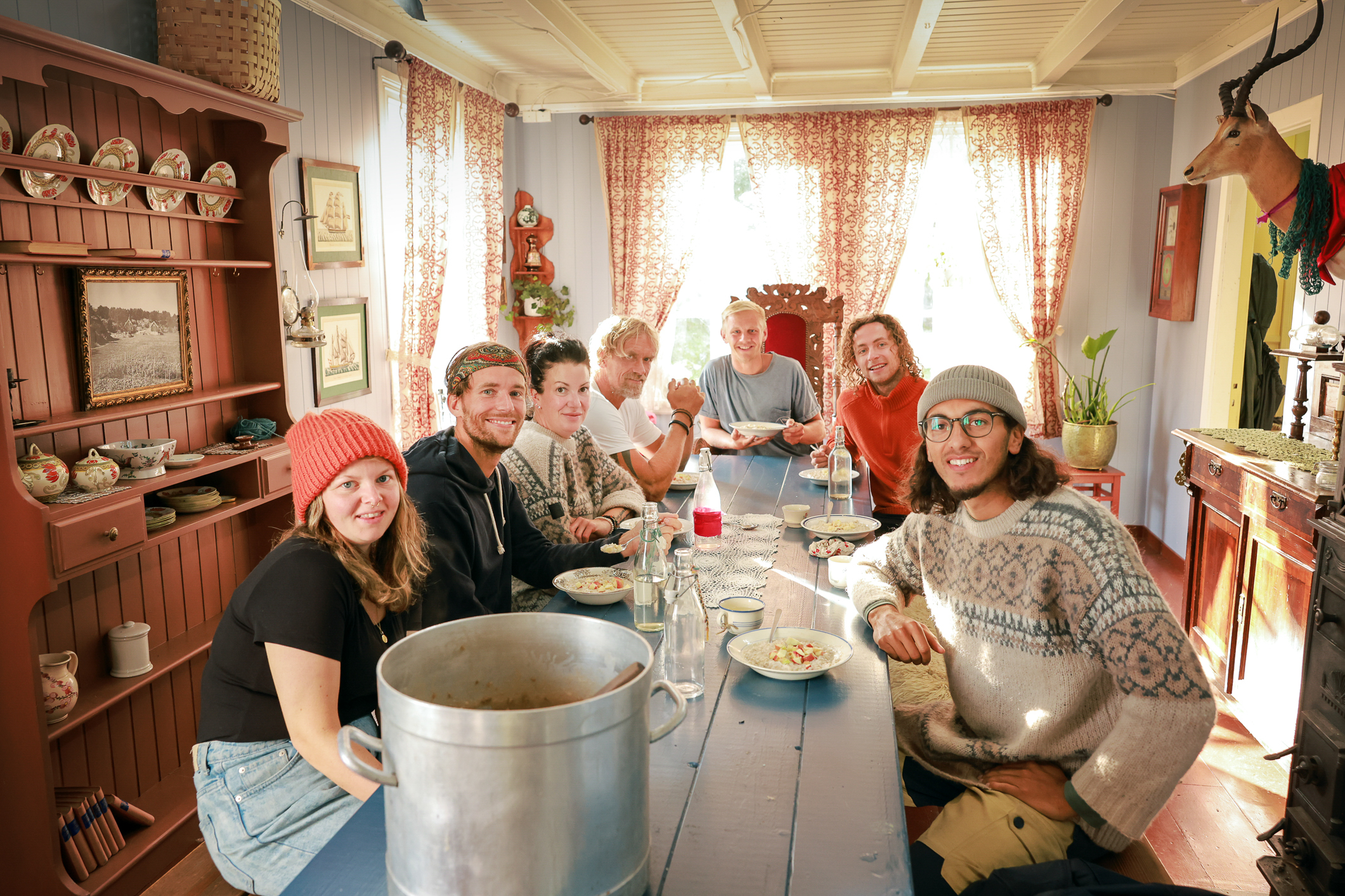 En gjeng med unge mennesker i avslappede klær rundt et måltid ved bordet. Stua de sitter i er fra en svunnen tid.