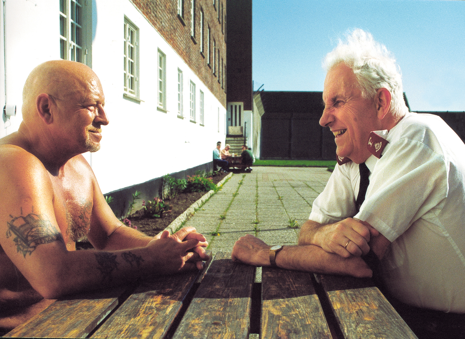 Bildet viser  Alv Tollisen som sitter ved et bord utendørs og snakker med en innsatt. Begge smiler, Tollisen har på seg uniformsskjorte, den andre mannen har bar overkropp og flere tatoveringer,
