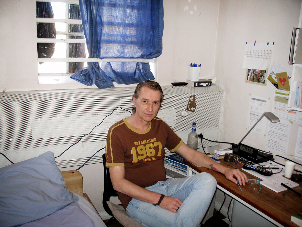 Bildet viser Sean som sitter på cella si på en stol mellom senga og skrivebordet. Bak ham er et vindu med gitter og blå gardin.