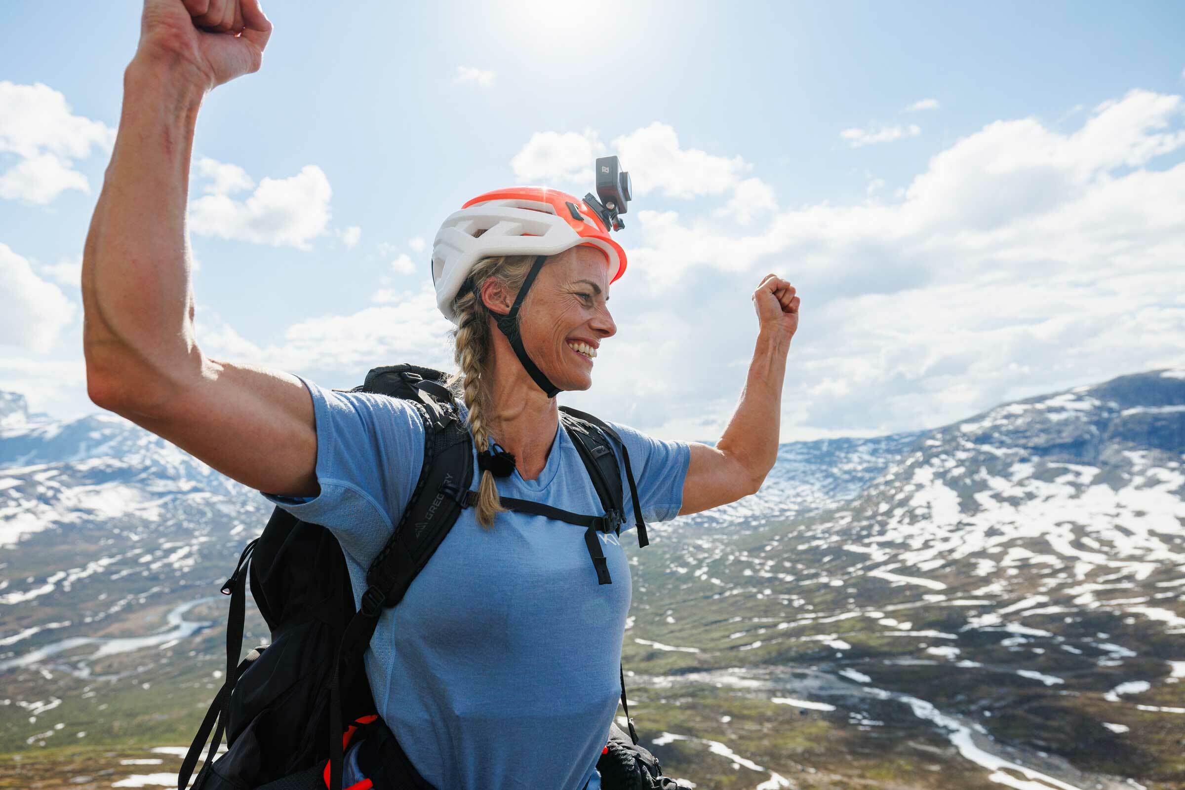 Kvinne med hjelm og ryggsekk i fjellheimen holder hendene i været.