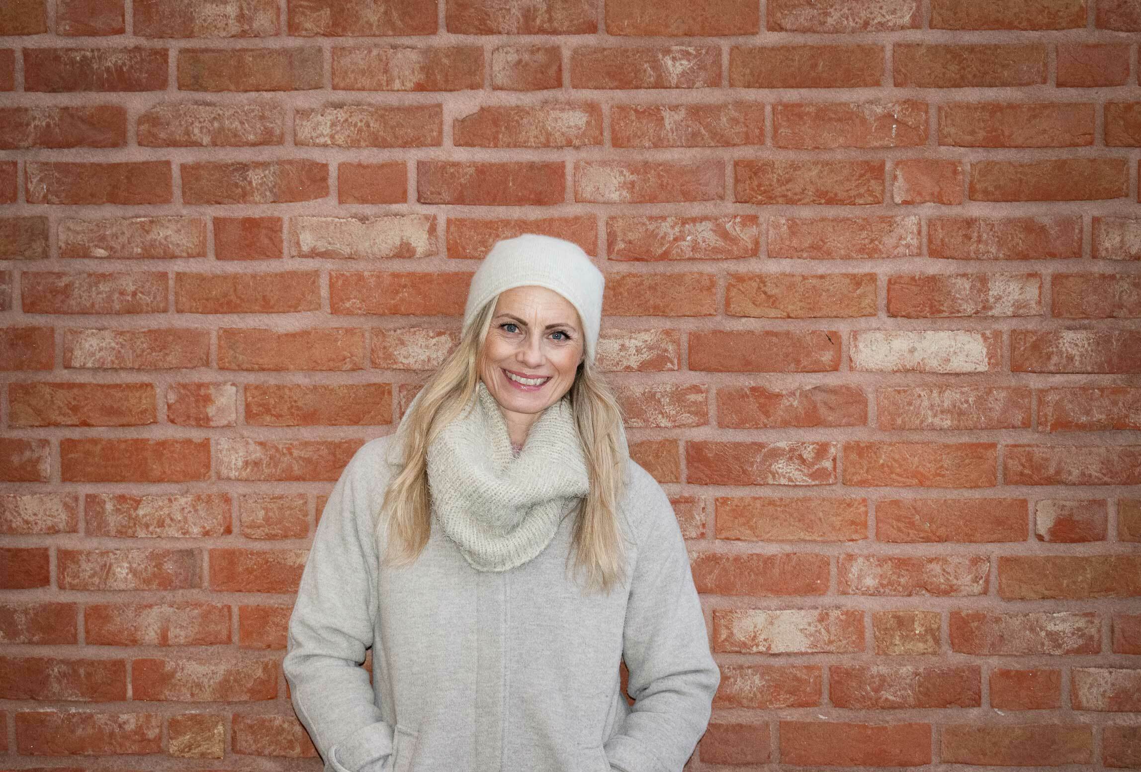 Kvinne med hvit lue og lys kåpe mot en rød murvegg.