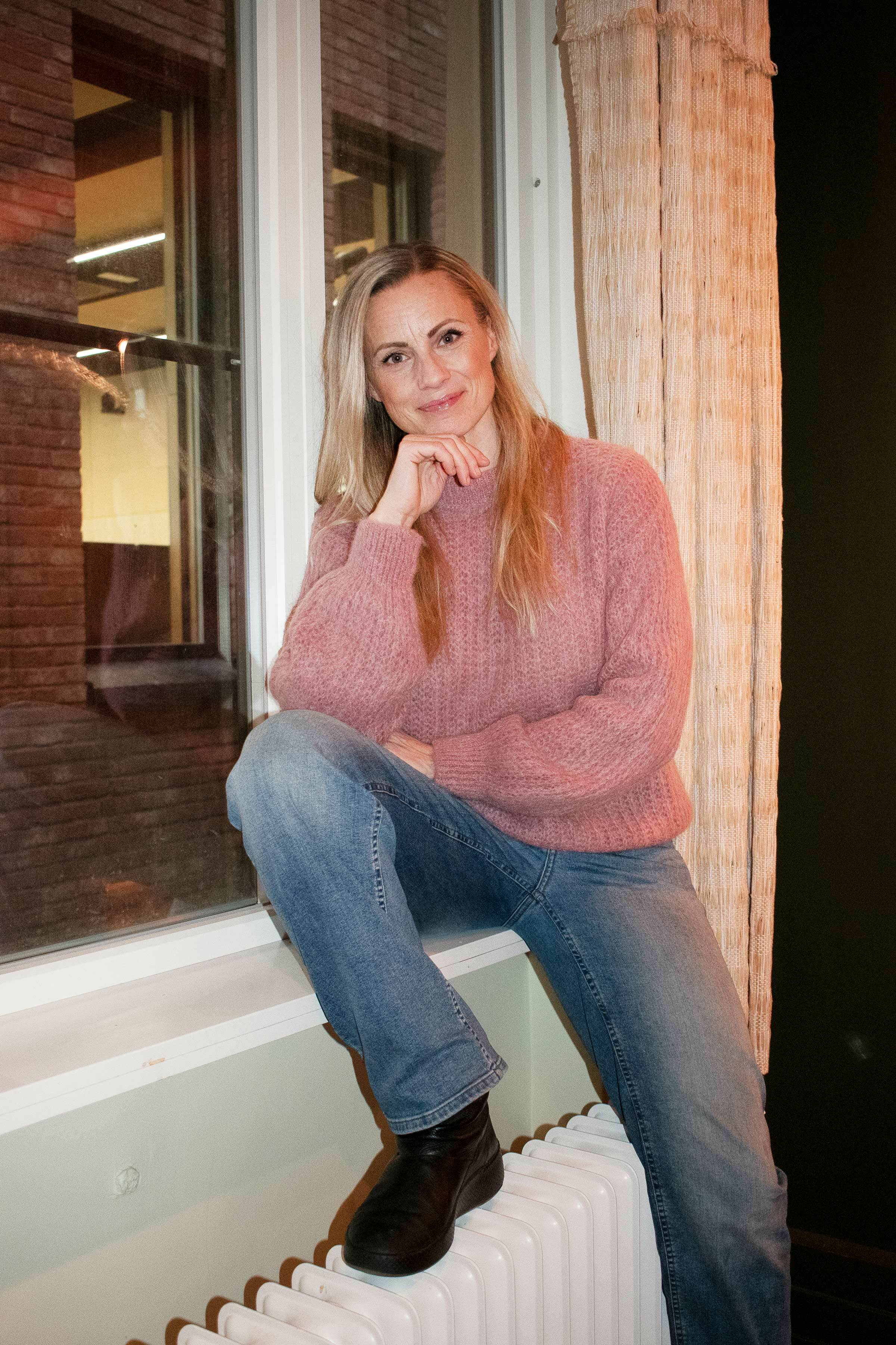 Kvinne  med rosa genser sitter i en vinduskarm med foten på en radiator.