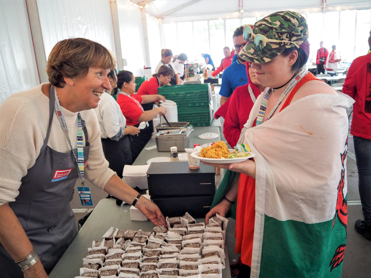 Foto: Voksen kvinne serverer mat til en av spillerne i et telt.