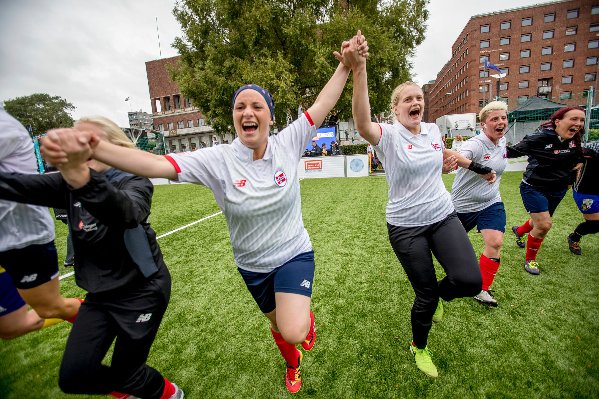 Foto: Lang rekke meg kvinnelige fotballspillerer holder hverandre i hendene mens de løper framover. I bakgrunnen sees rådhuset i Oslo. Tre av dem har på seg hvite spillertrøyer, de andre blå.