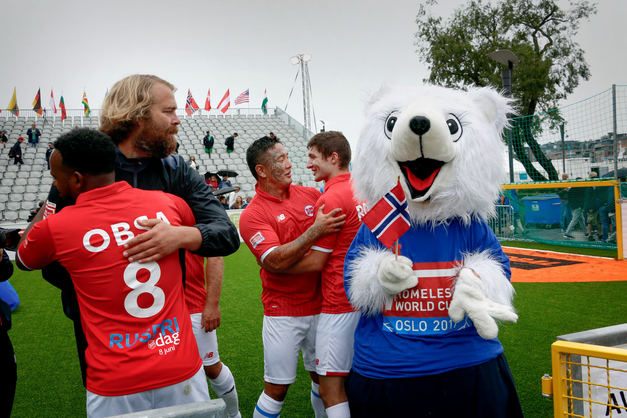 Foto: Menn i norske, røde fotballdrakter som klemmer hverandre. I bakgrunnen er det store tribuner. På banen står også en maskot, en hvit isbjørn med blår trøye det det står Homeless World Cup Oslo 2017. Han holder et norsk flagg.