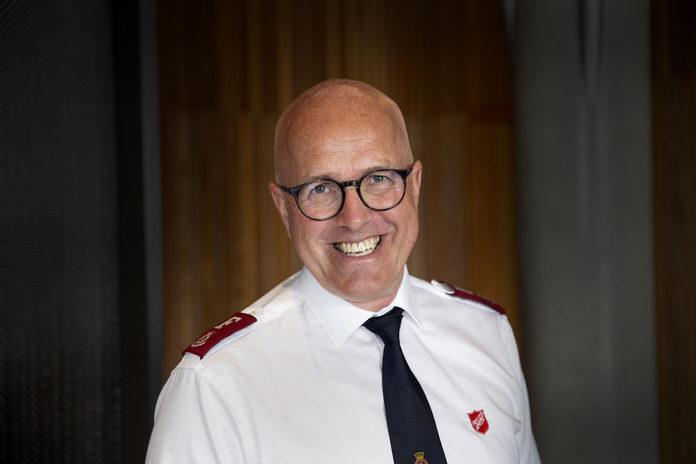 Foto: Mann med glattbarbert hode, runde briller og et stort smil ser rett i kamera. Han har på seg hvit uniformstrøye fra Frelsesarmeen og mørkt slips.