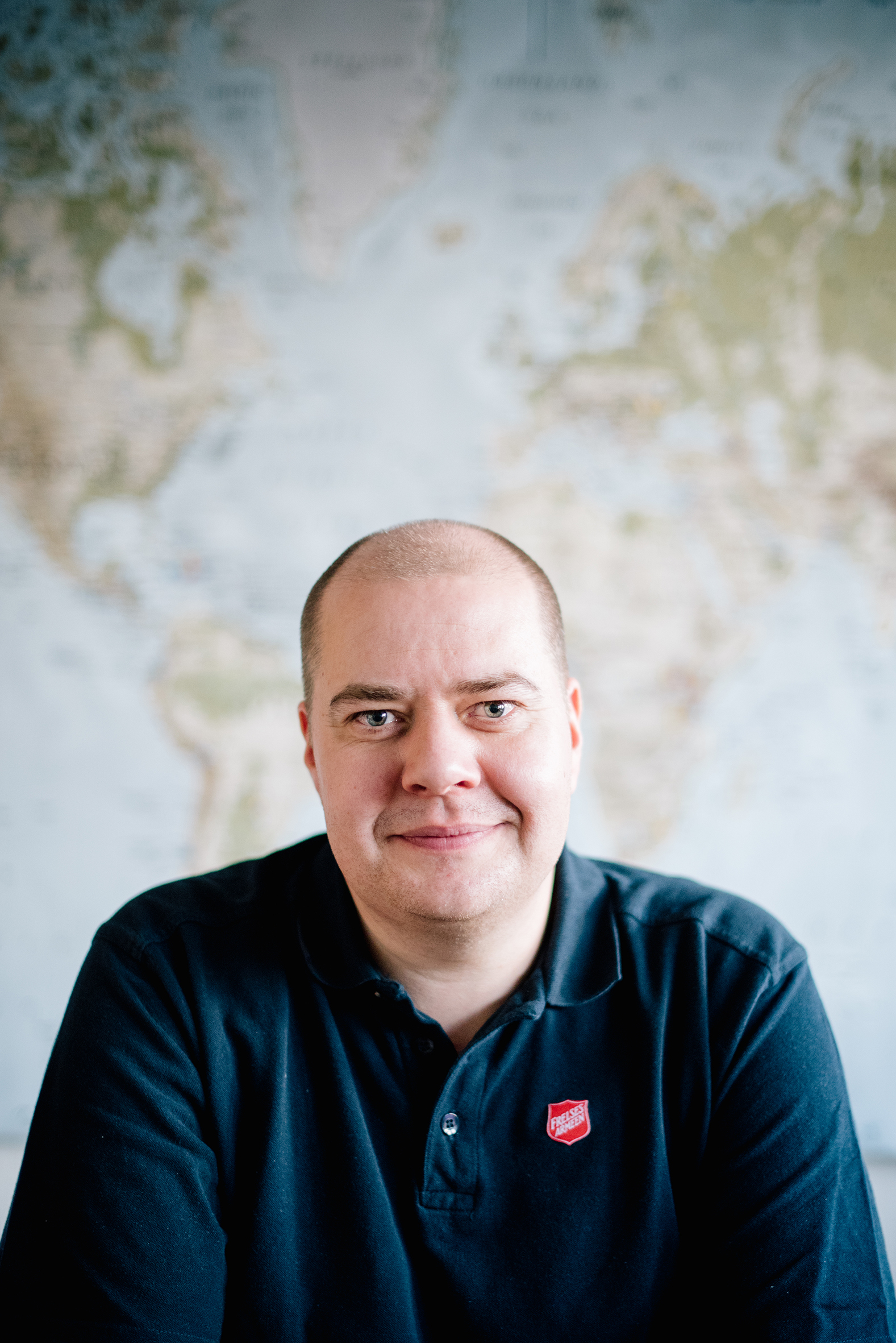 Foto: Mann i 40-årene med mørkt hår som er tynt på toppen. Han har på seg mørk blå skjorte med rød logo fra Frelsesarmeen. Bak ham er et kart over hele verden.