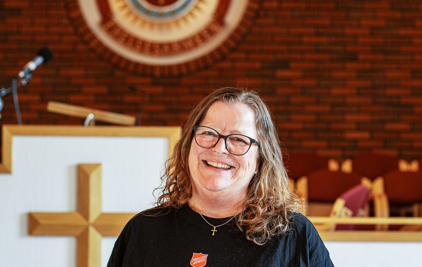 Middelaldrende kvinne med briller og halvlangt hår står foran en prekestol med Frelsesarmeens skjold på t-skjorten.