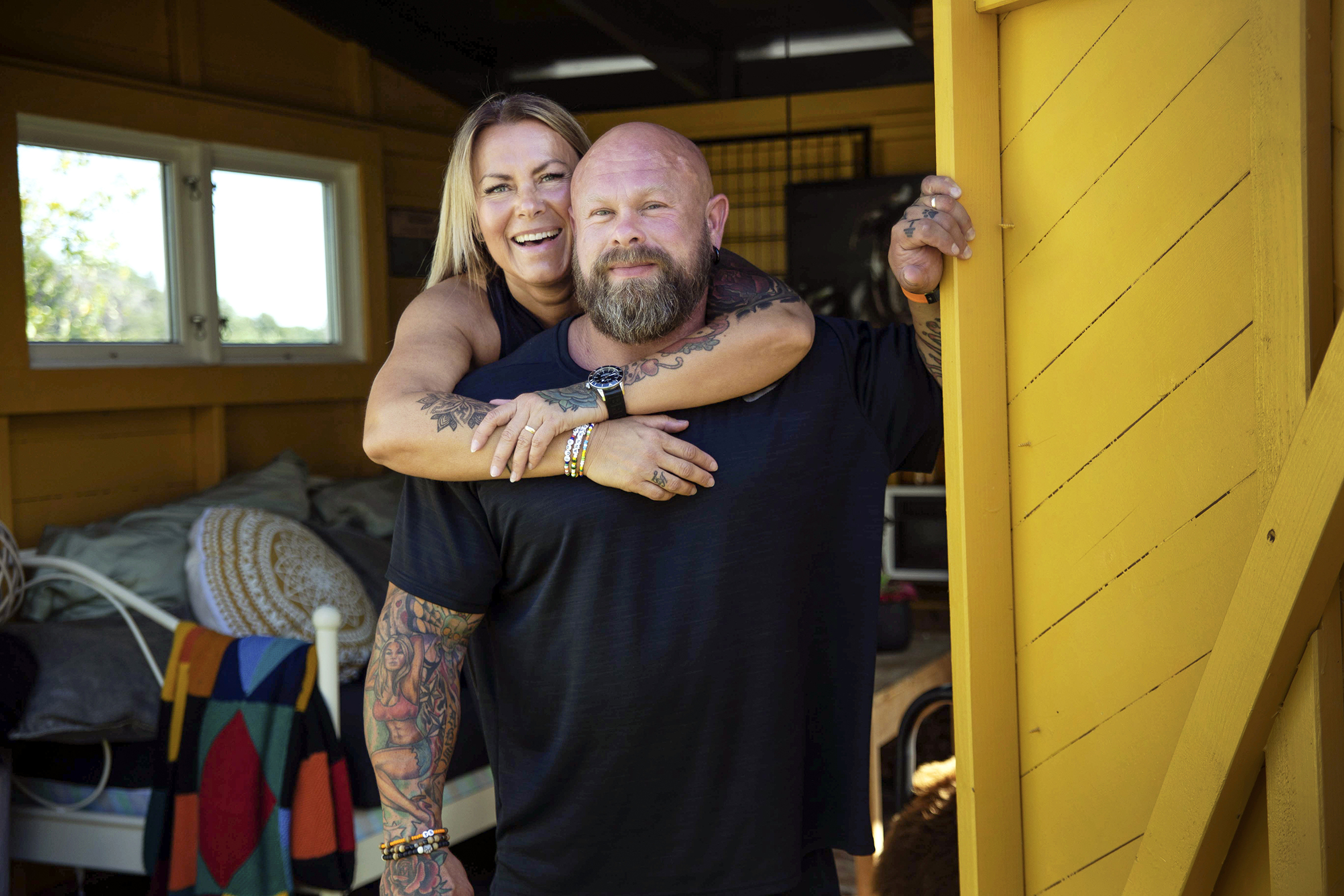 Foto: Viser kvinne med tatoveringer som henger på ryggen bak en muskuløs mann – også han med tatoveringer. De står i døra inn til et gjesterom med separat inngang.