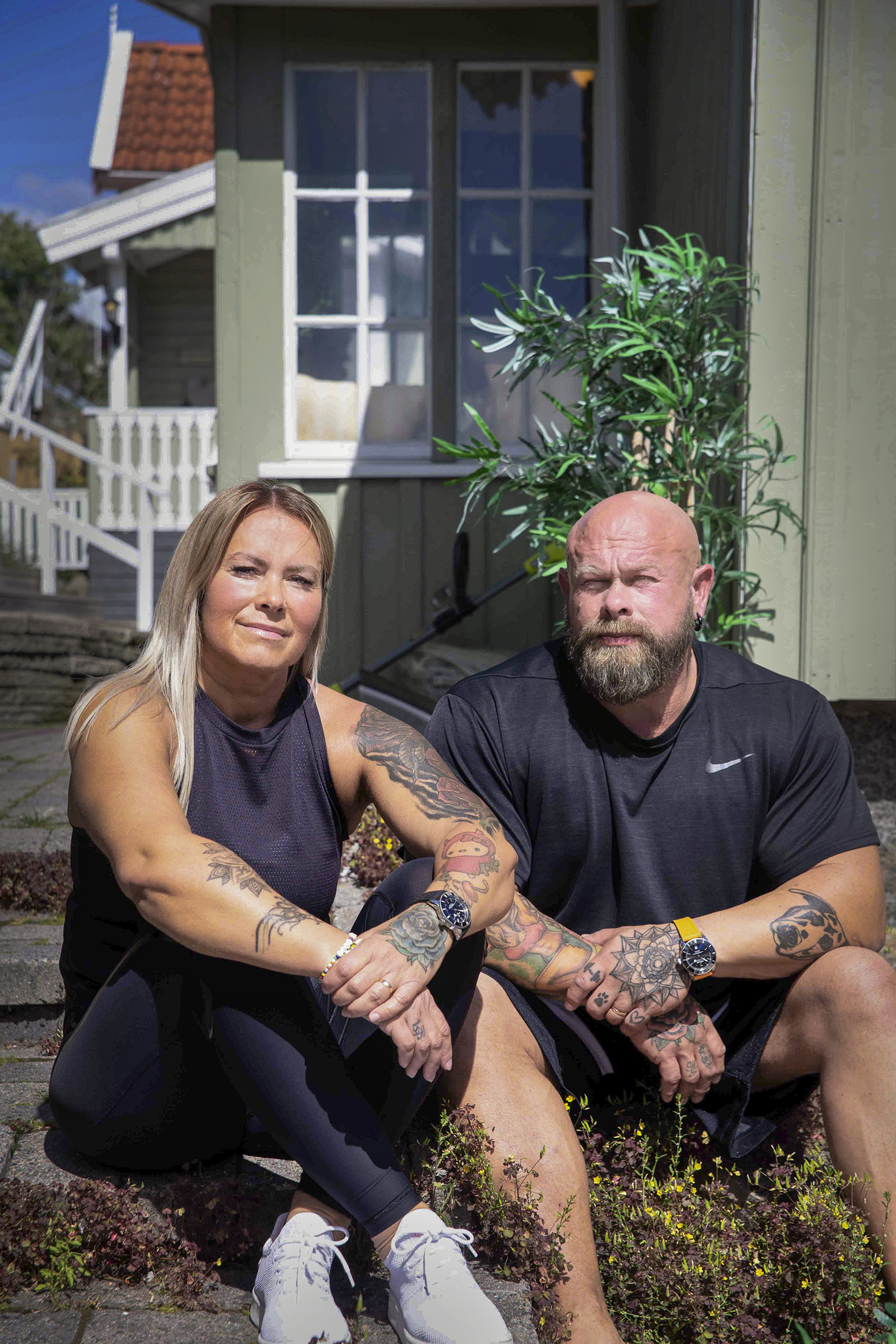 Foto: Kvinne om mann – begge med mange tatoveringer på armene – sitter solbrune i en trapp utenfor et hus.