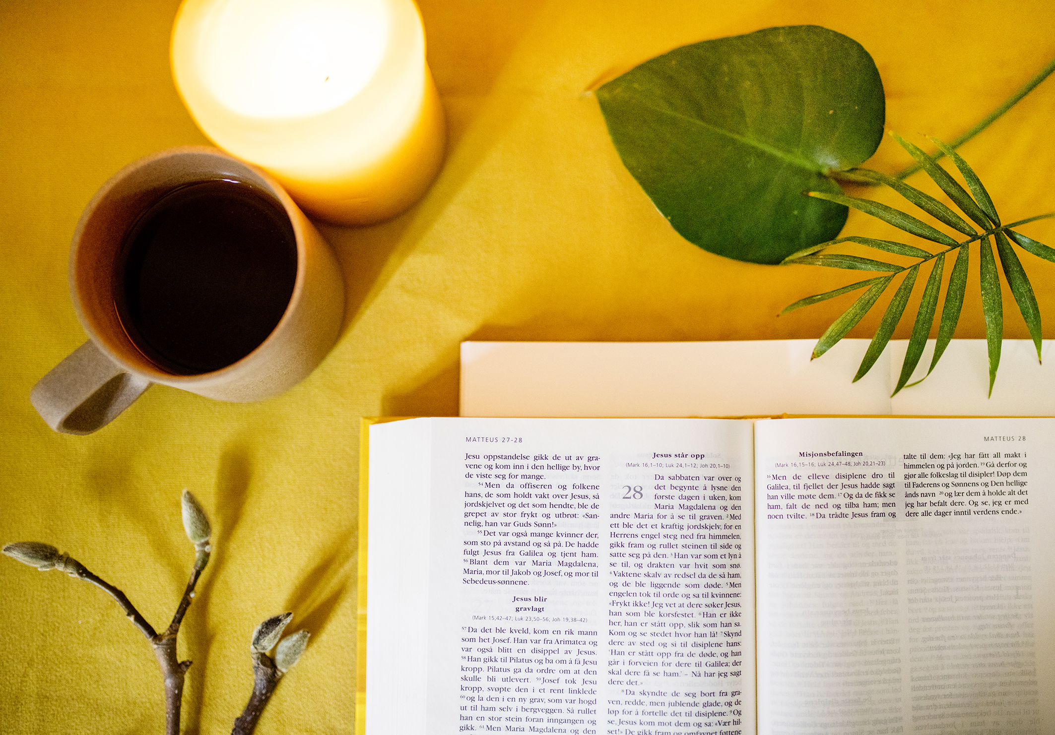 Bilde av en oppslått bibel, en kaffekopp, et tent lys og noen grønne blader. Alt på en gul duk.