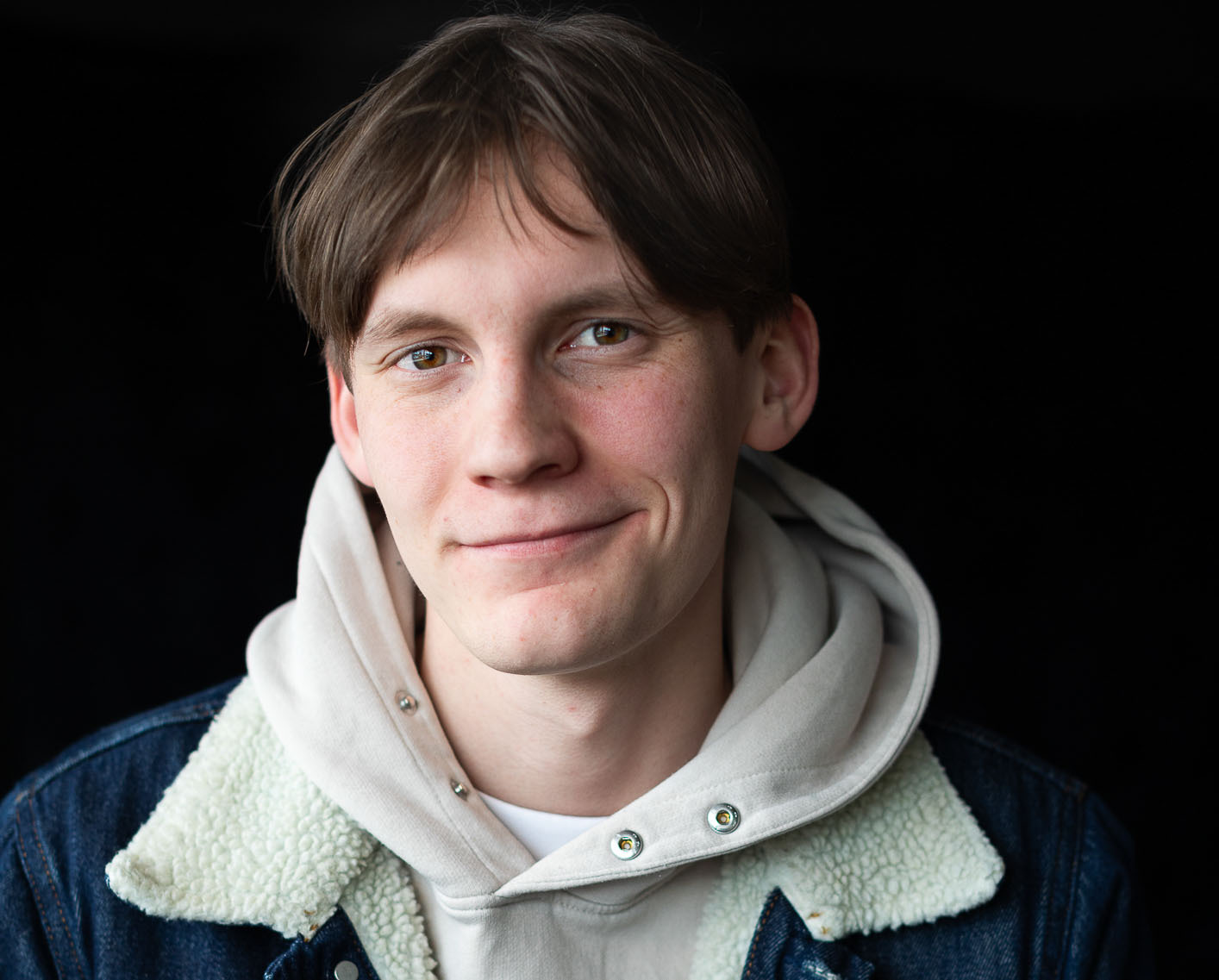 En ung gutt med brunt hår og dongerijakke smiler.