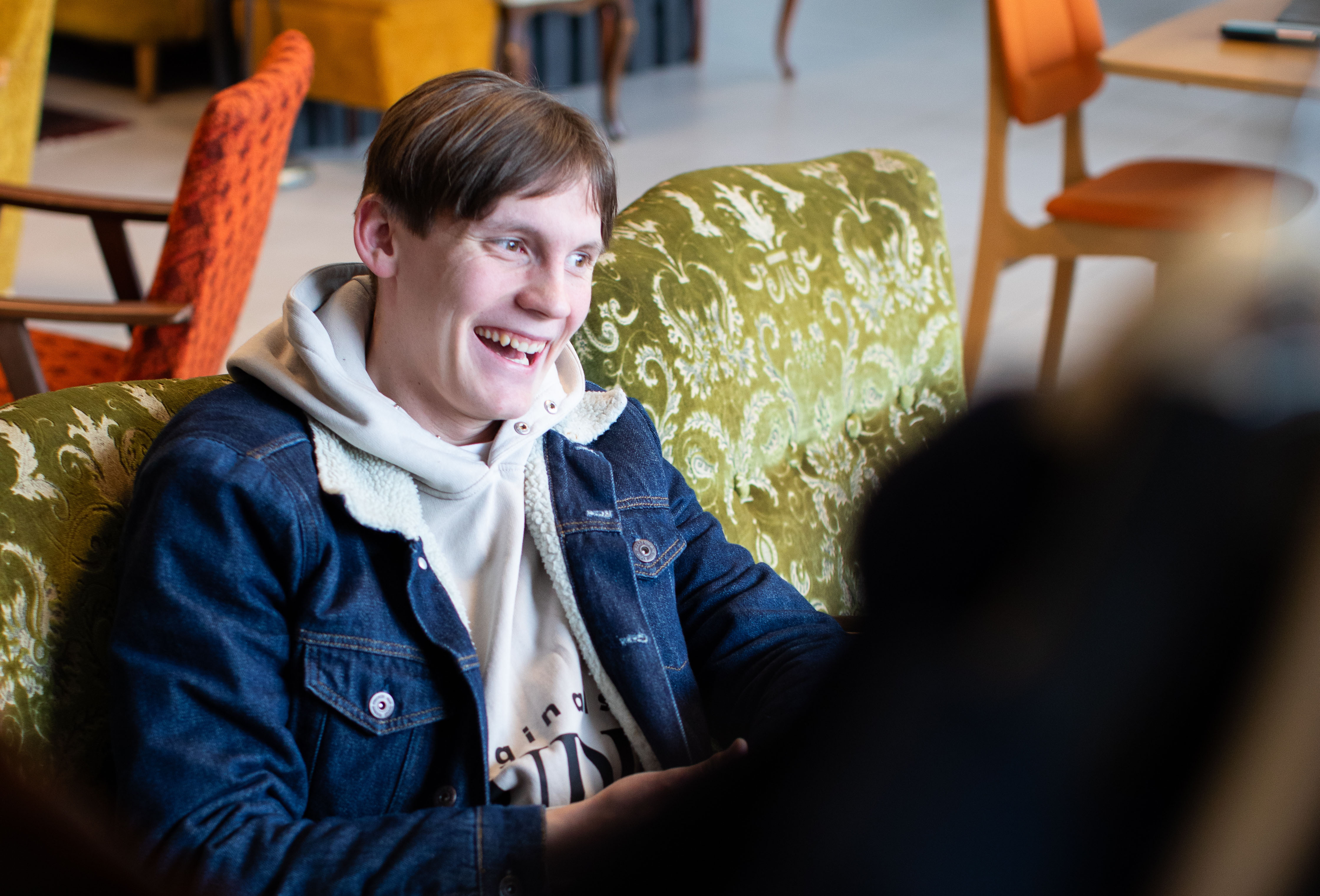 En ung gutt med brunt hår og dongerijakke ler og prater med en annen i korpset. Vi ser gamle møbler i bakgrunnen.
