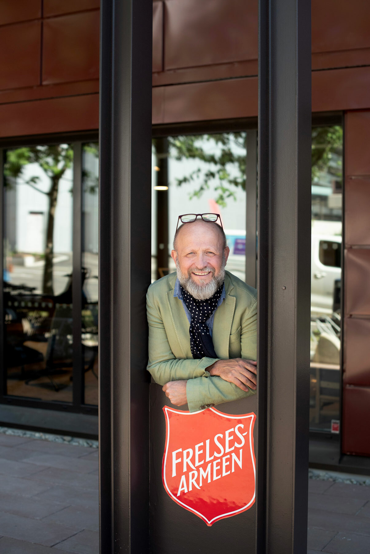 Foto: Mann i lys grønn dressjakke og prikkete tørkle i halsen står rett over et rødt skilt med logoen til Frelsesarmeen. Han har skjegg og briller på et hode uten hår.