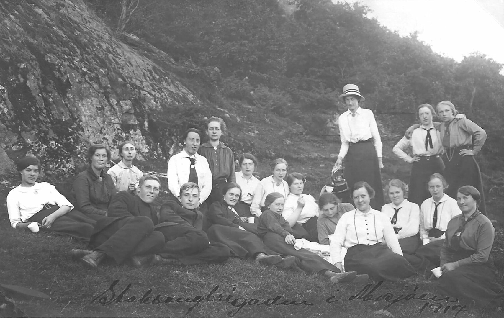 Foto: Gammelt svart-hvitt-bilde av unge kvinner og menn i datidens uniformer. De står, sitter og ligger i en utmark med fjell like bak.