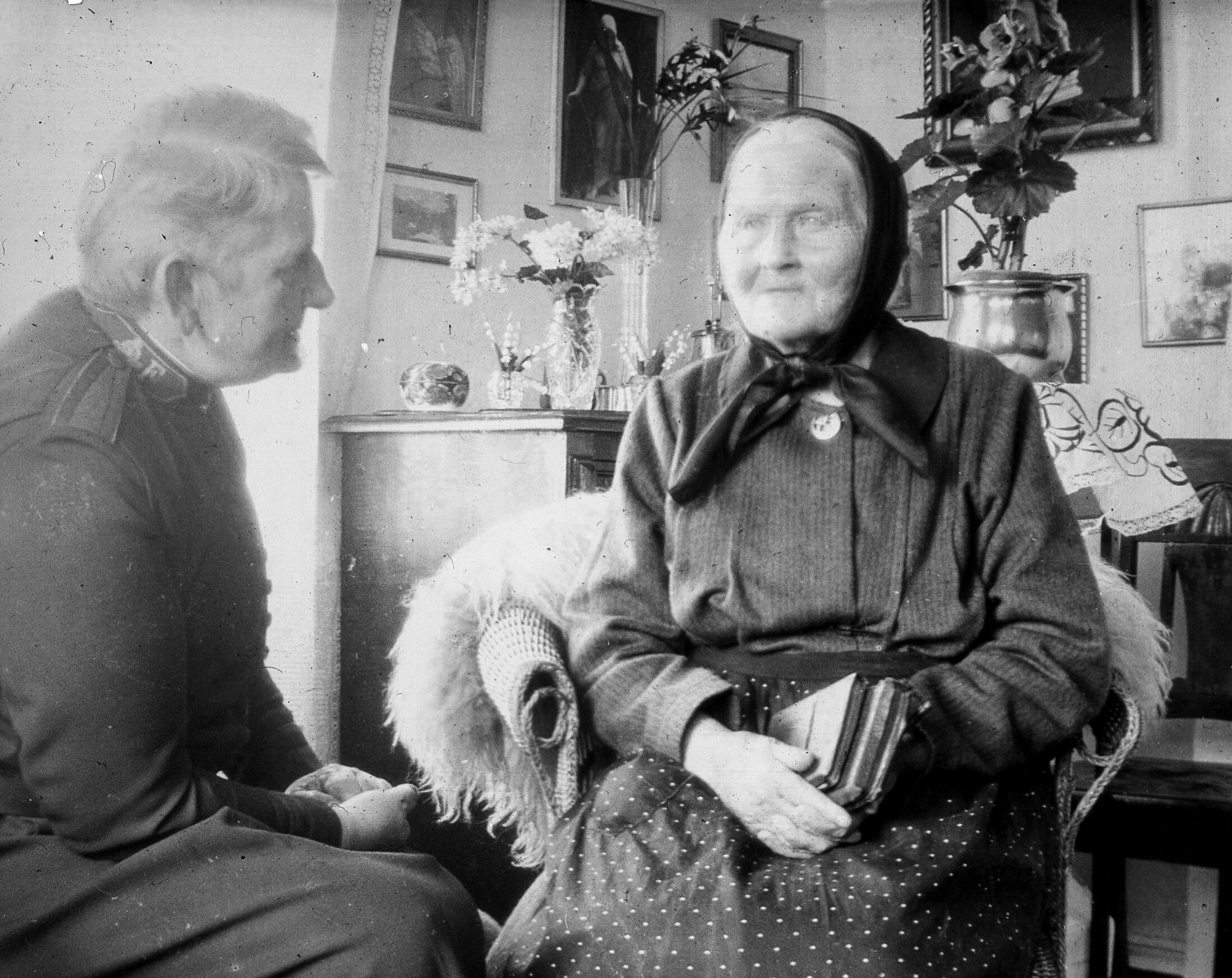 Foto: Gammelt svart-hvitt-bilde av en kvinne med kortklippet hår i en uniform fra Frelsesarmeen. Hun snakker med en gammel dame med skaut på hodet i et hjemmekoselig rom med bilder på veggen bak.