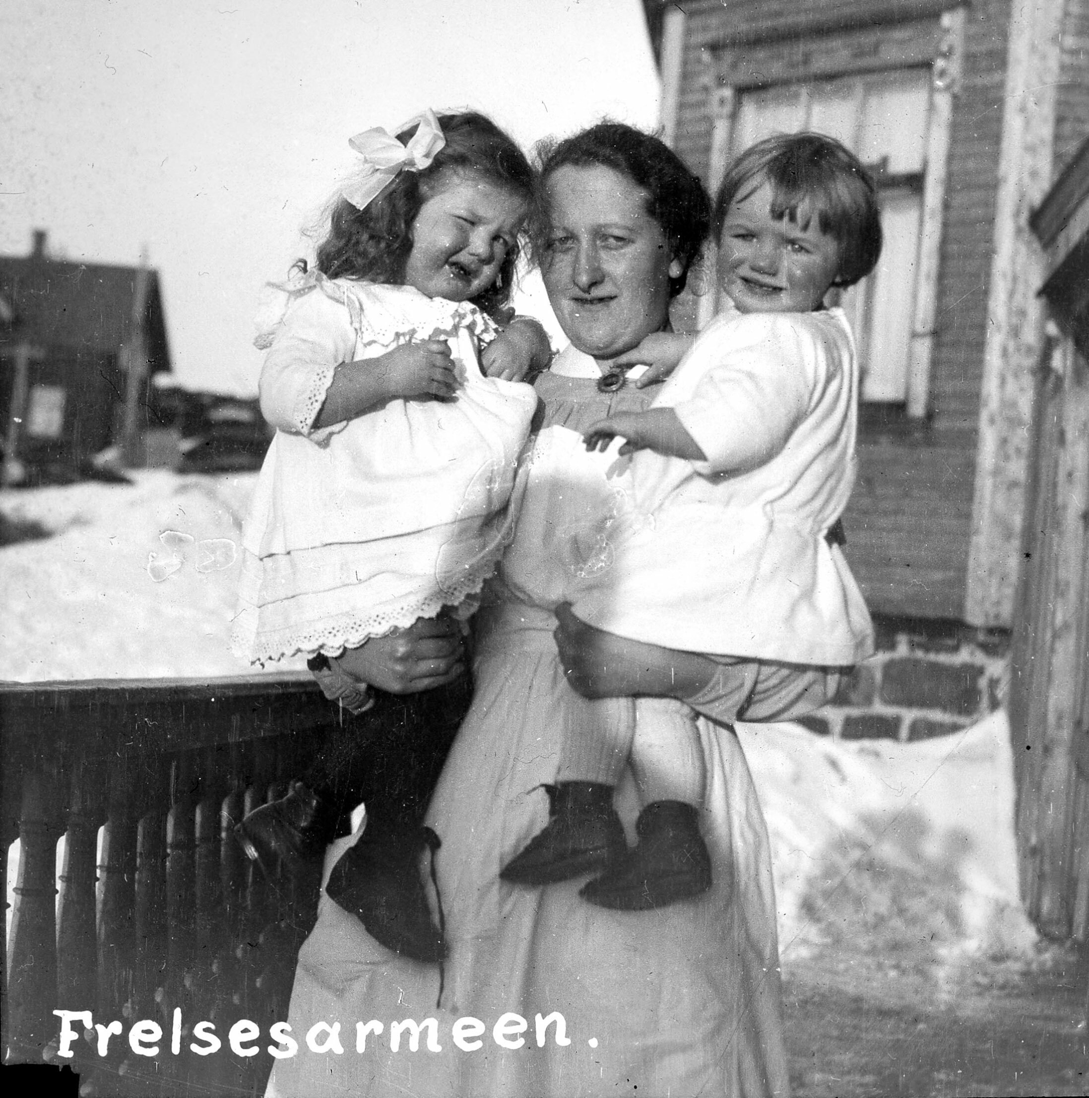 Foto: Gammelt svart-hvitt-bilde av en slumsøster i uniform som bærer to pent kledde barn. Hun står utenfor et hus og det er rester av snø på bakken her og der.