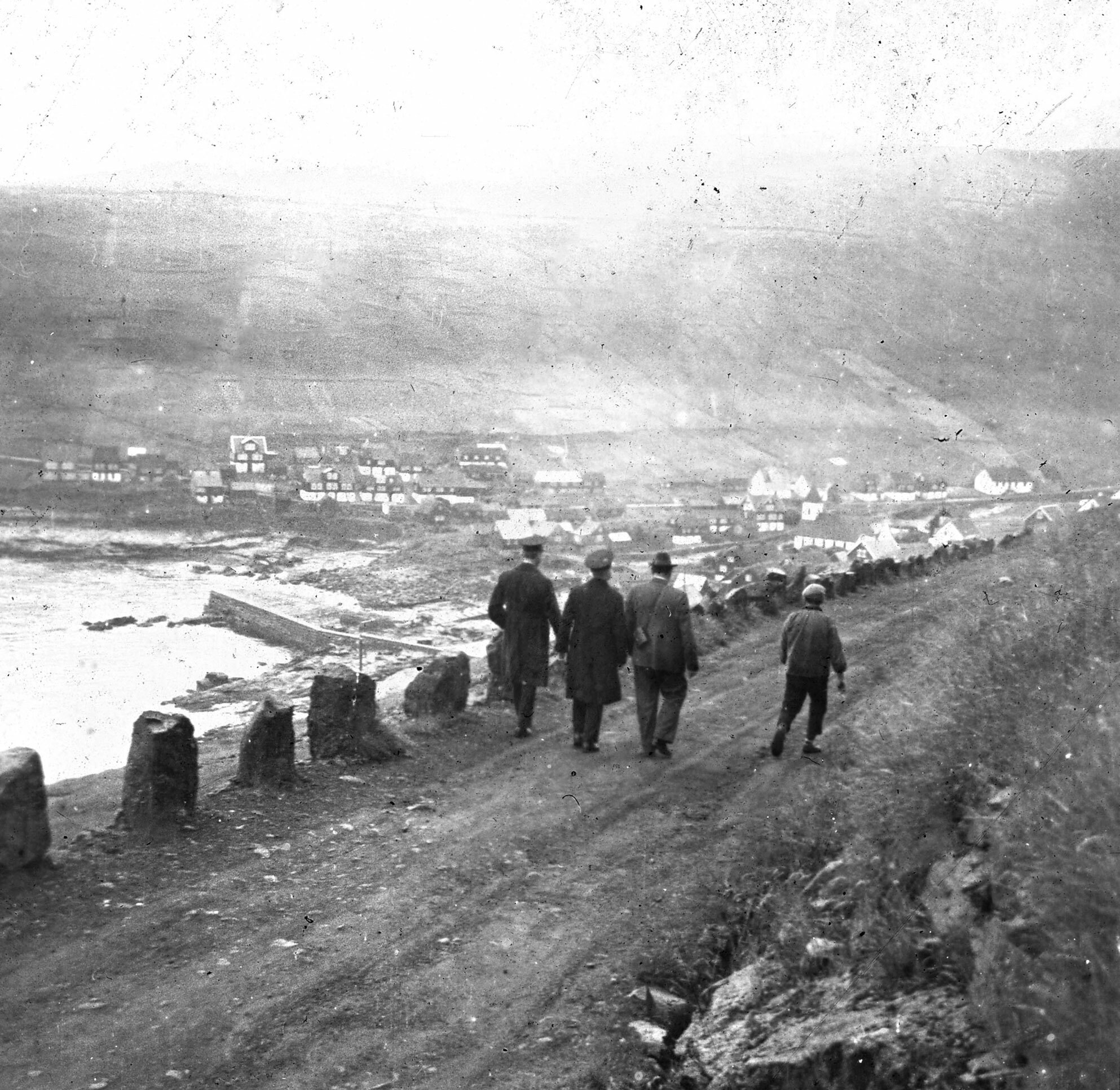 Foto: Gammelt bilde i svart hvitt av en ung gutt som går sammen med tre voksne menn, der to av dem er i uniform fra Frelsesarmeen. De går langs en grusvei, og lenger nede ligger noen få, små trehus ved sjøen.