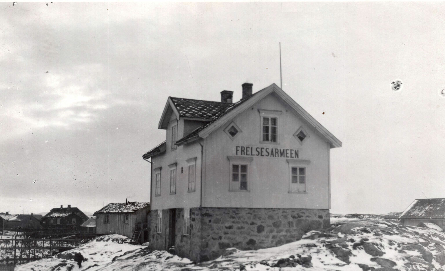Foto: Gammelt svart-hvitt-bilde med et hvitt hus i tre etasjer. Det står på et litt øde sted sammen med noen få hus til. Her er det ingen trær, men litt snø på bakken.
