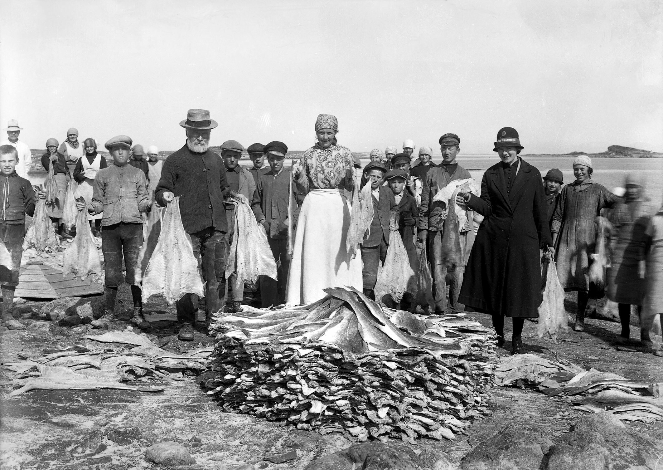 Foto: Gammelt svart-hvitt-bilde av en rekke folk som bærer klippfisk som de legger i en haug. Her deltar menn, kvinner og barn i arbeidet. det samme gjør em kvinne i uniform med hatt.