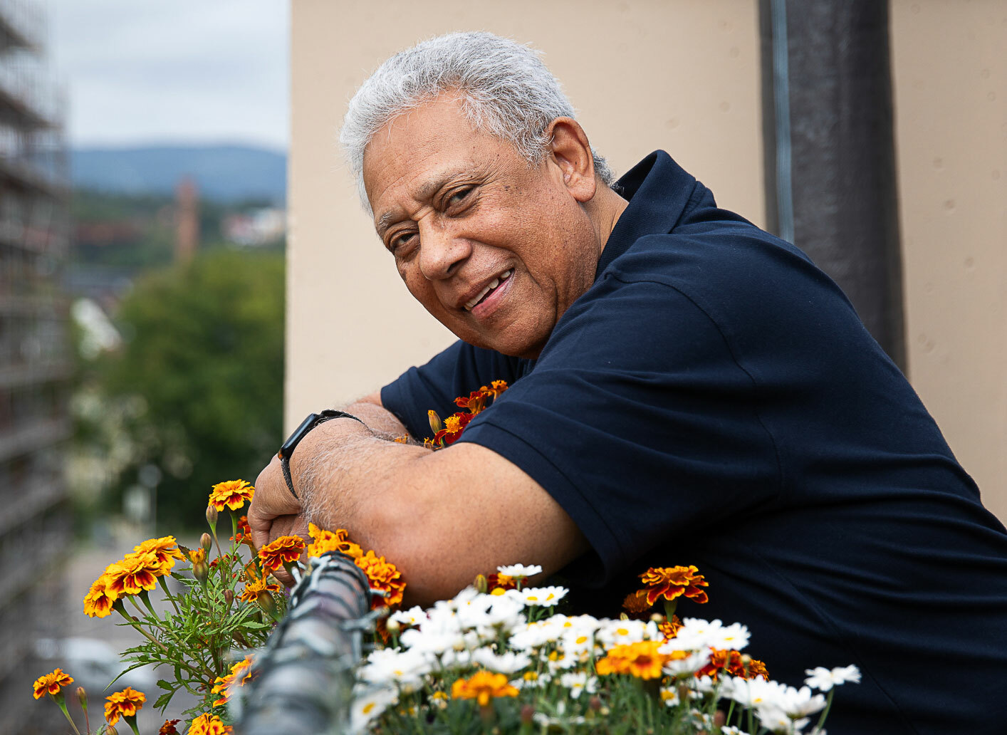 Mann med grått hår står bøyd over en blomsterkasse på terassen sin i ei blokk. Velstelt og mange fine planter.