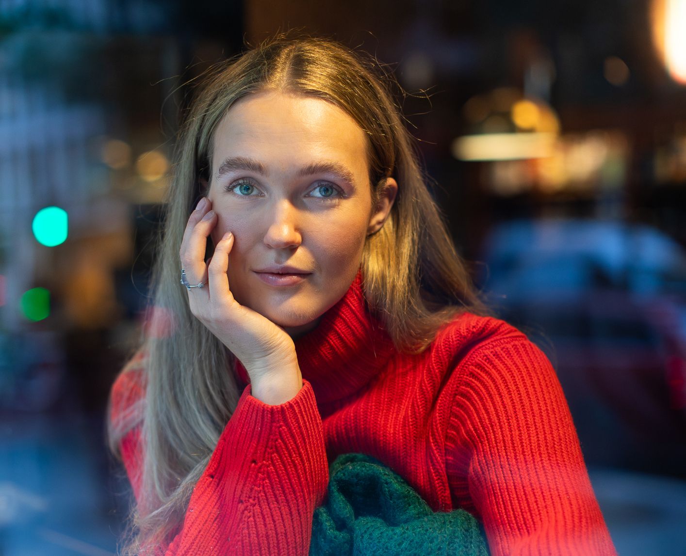 Ung kvinne med langt hår fotograferes inn gjennom et kafé-vindu. Hun har på seg julerød genser og lener hodet på sin høyre hånd.