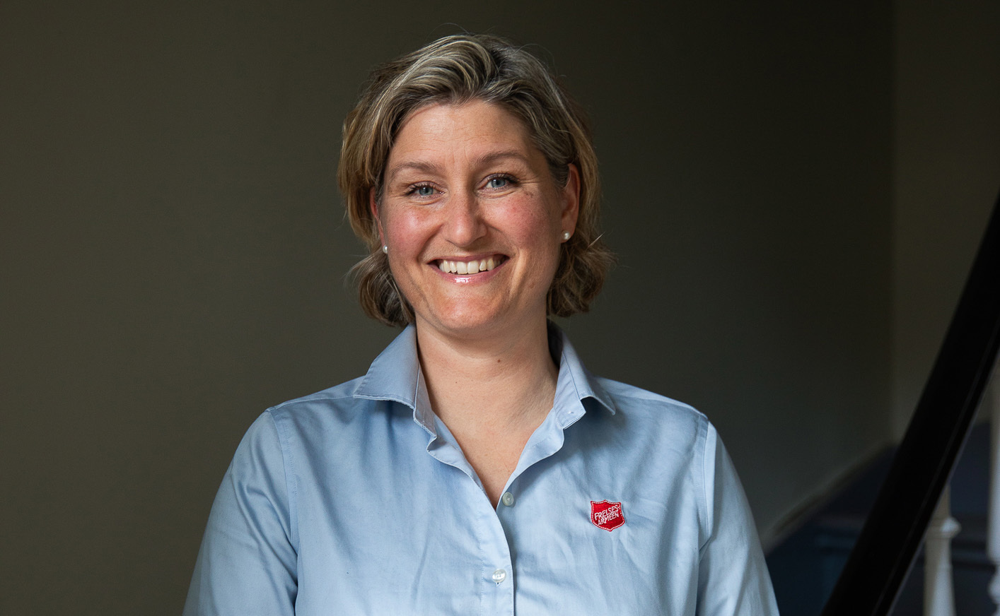 Smilende kvinne med lyseblå skjorte med Frelsesarmeens røde skjold på brystet.