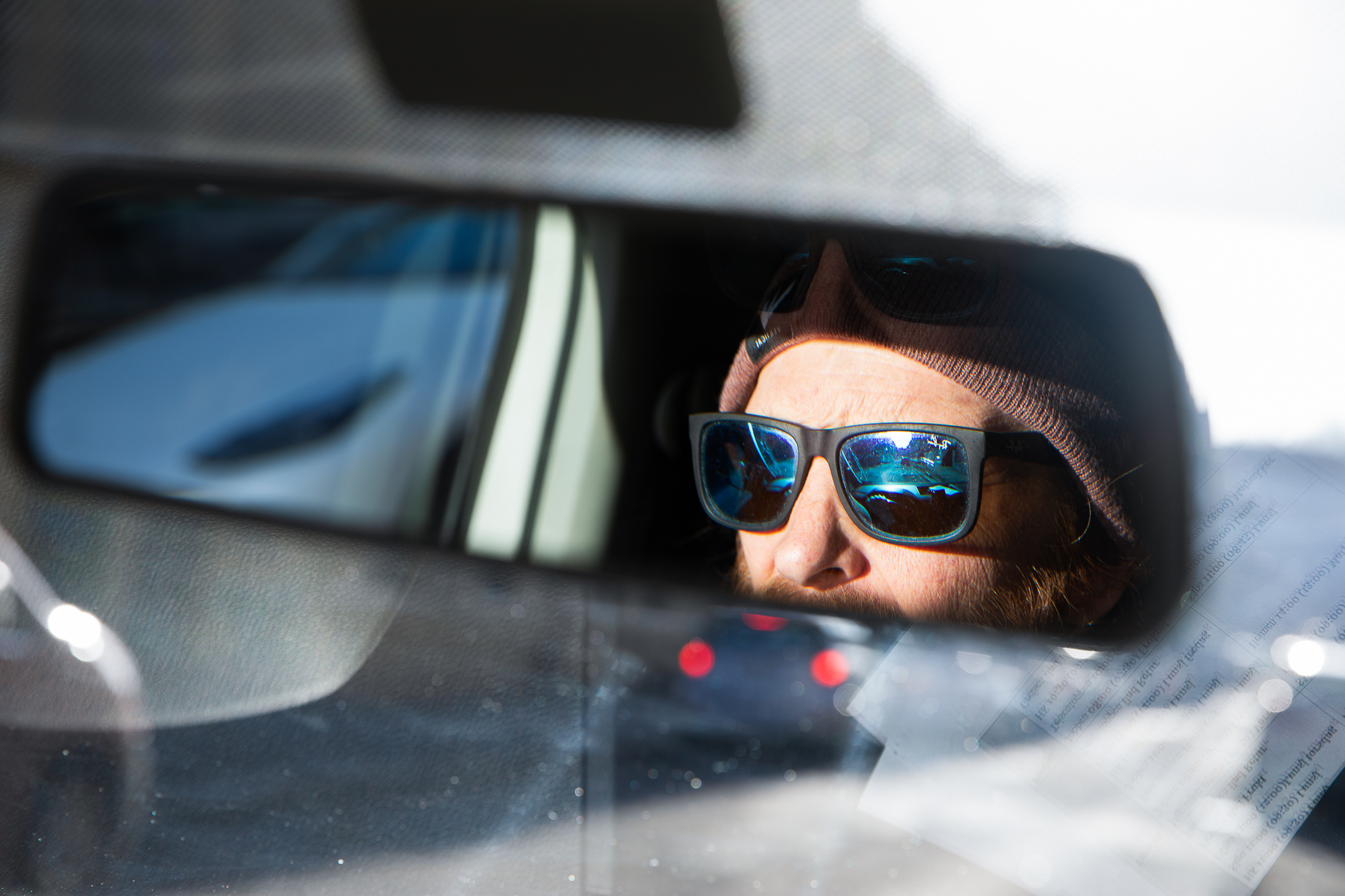 Mann med solbriller og lue ser i et bilspeil.