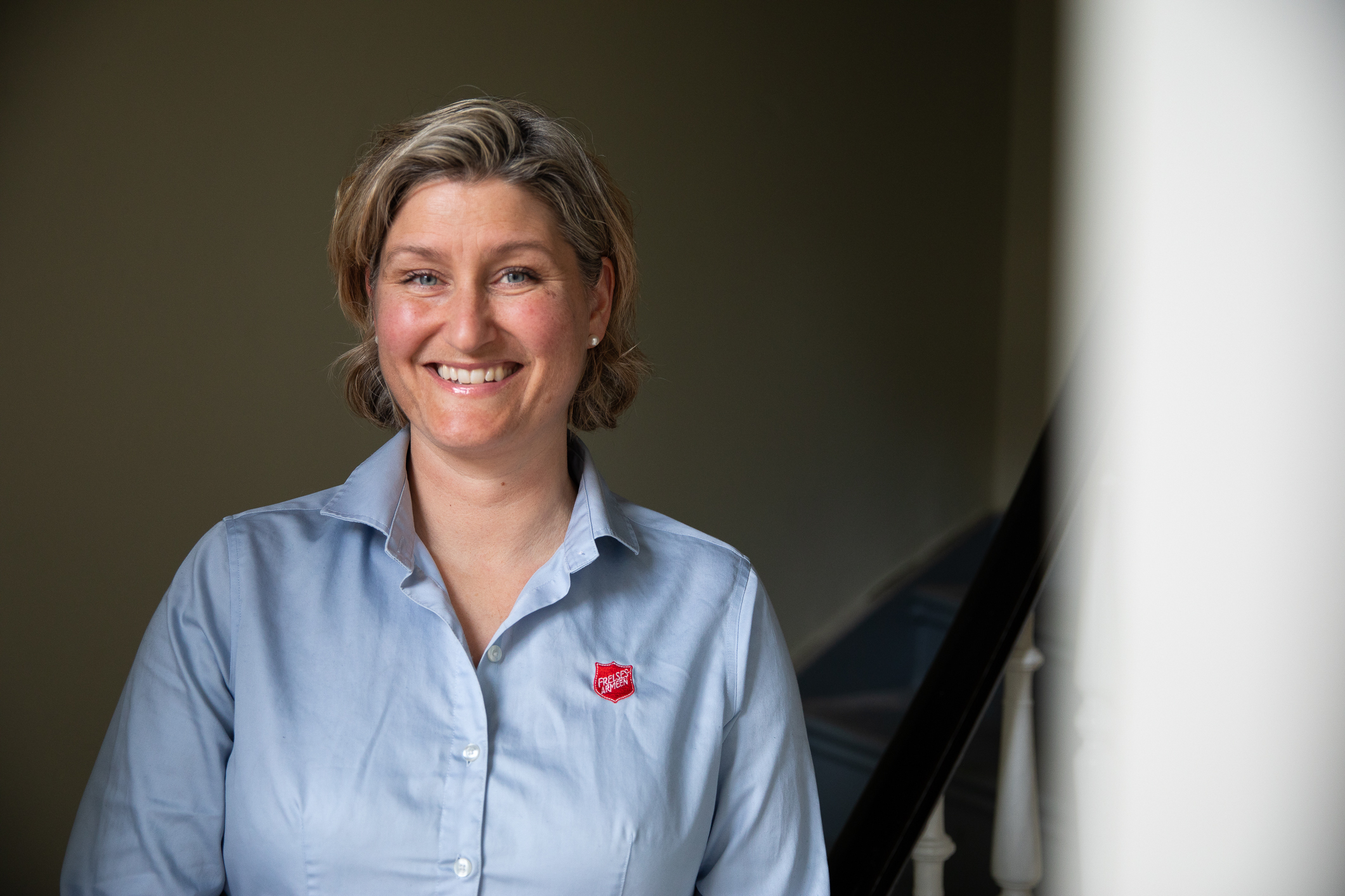 Smilende kvinne med lyseblå skjorte med Frelsesarmeens røde skjold på brystet.
