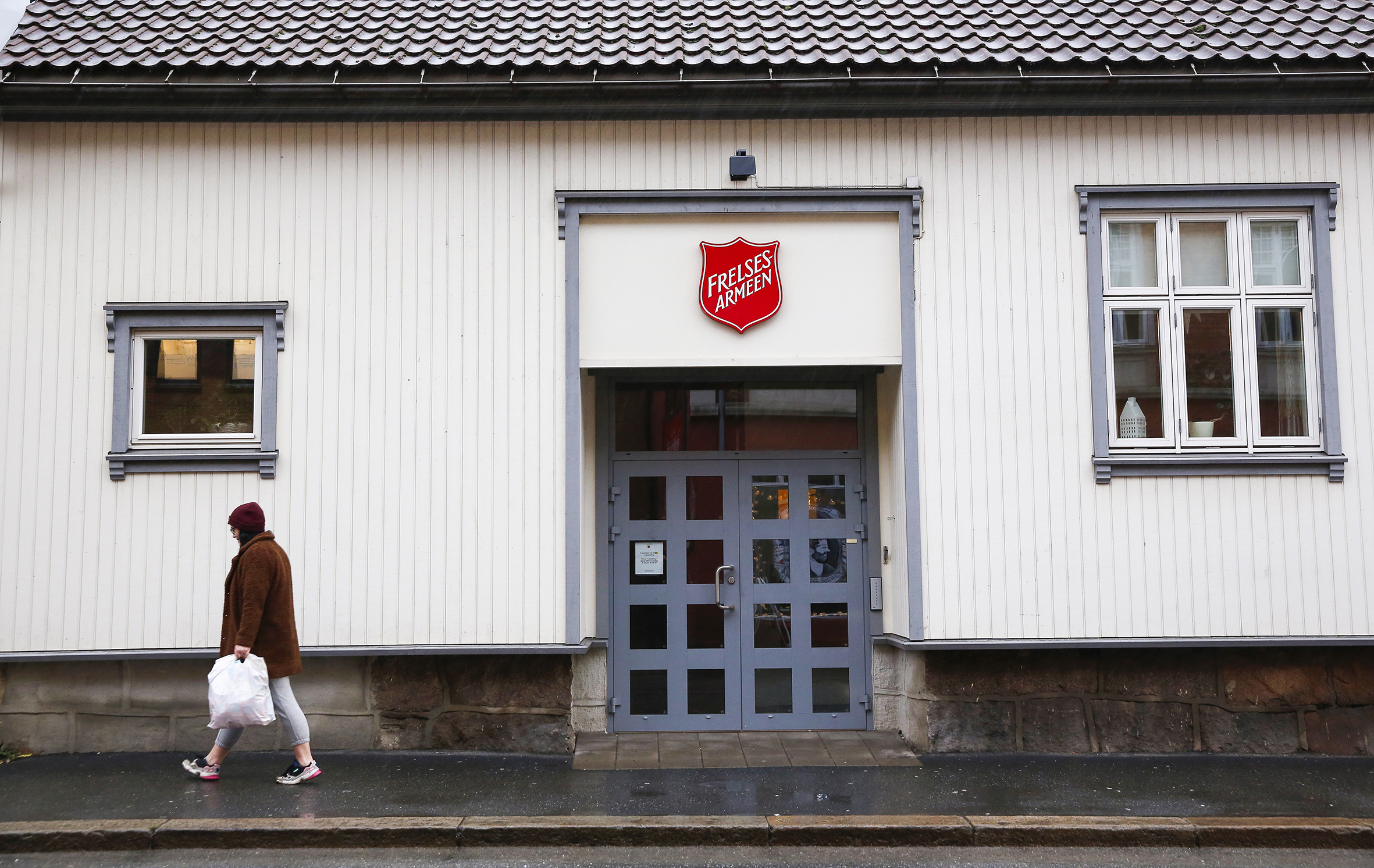 Bilde av et bygg med Frelsesarmeens logo utenpå. En kvinne passerer med hette over hodet og en pose i hånden.