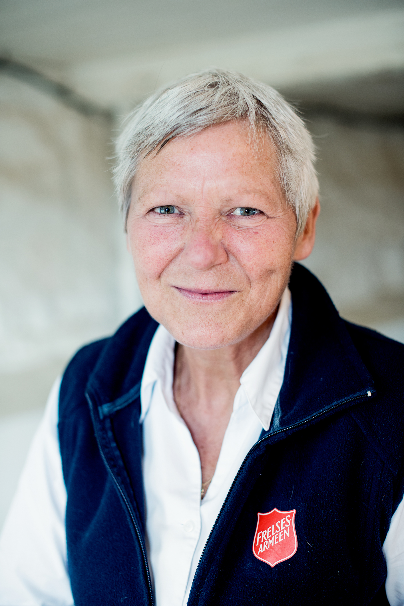 Voksen kvinne ser varmt inn i kamera. Har på seg hvit skjorte og blå vest med logo fra Frelsesarmeen.