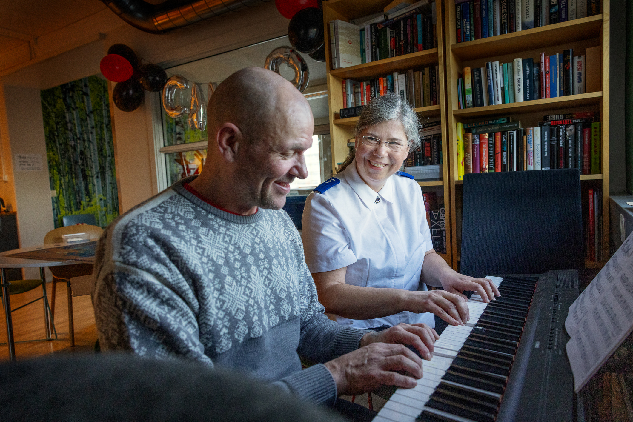 Voksen mann i strikkegenser og kvinne uniformsskjorte fra Frelsesarmeen sitter sammen ved et piano og spiller side om side.