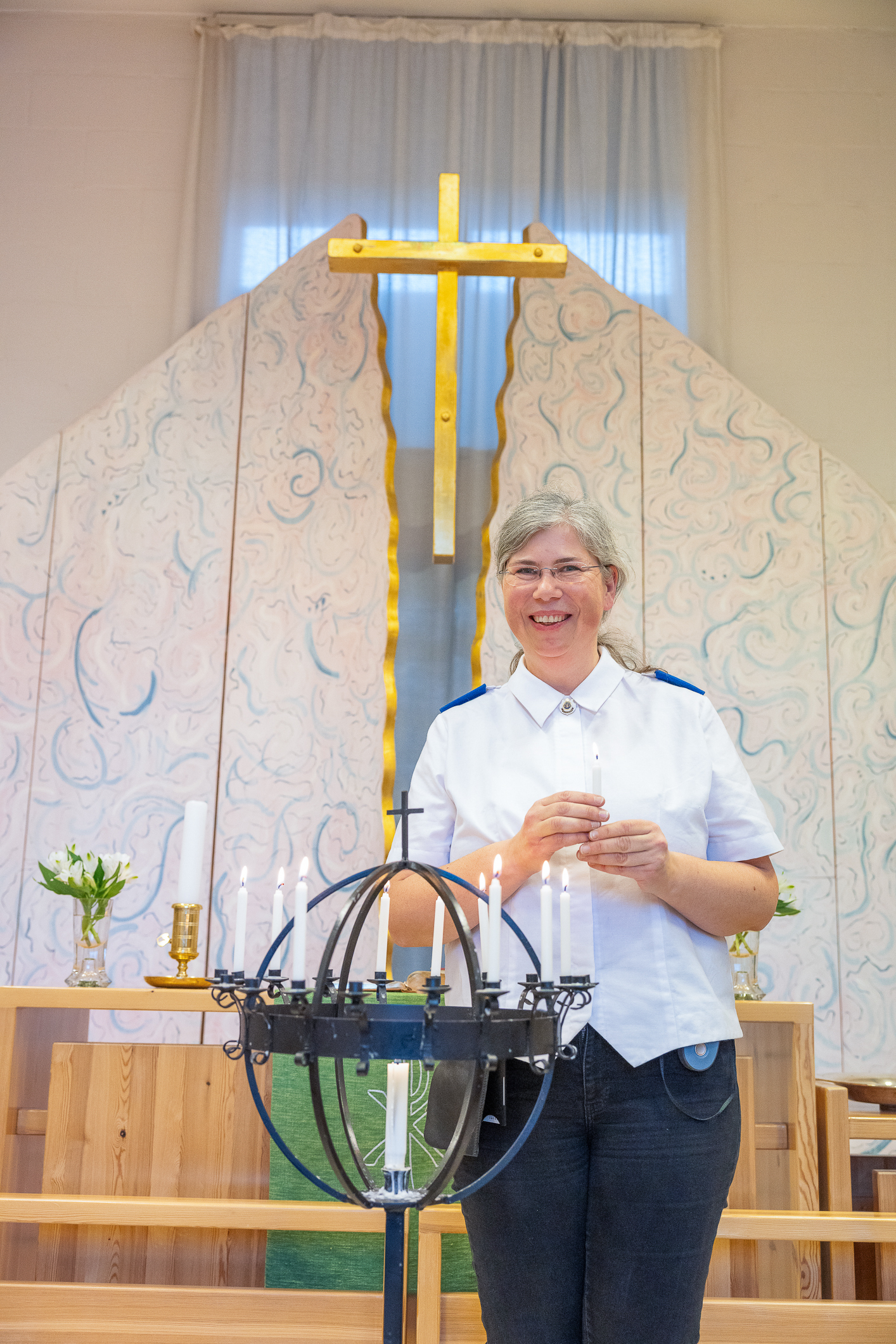 Kvinne i uniformsskjorte tenner lys i et kirkerom. Bak på veggen henger et kors og lysestaken i smijern er formet som en globus og står på et høyt stativ på gulvet.