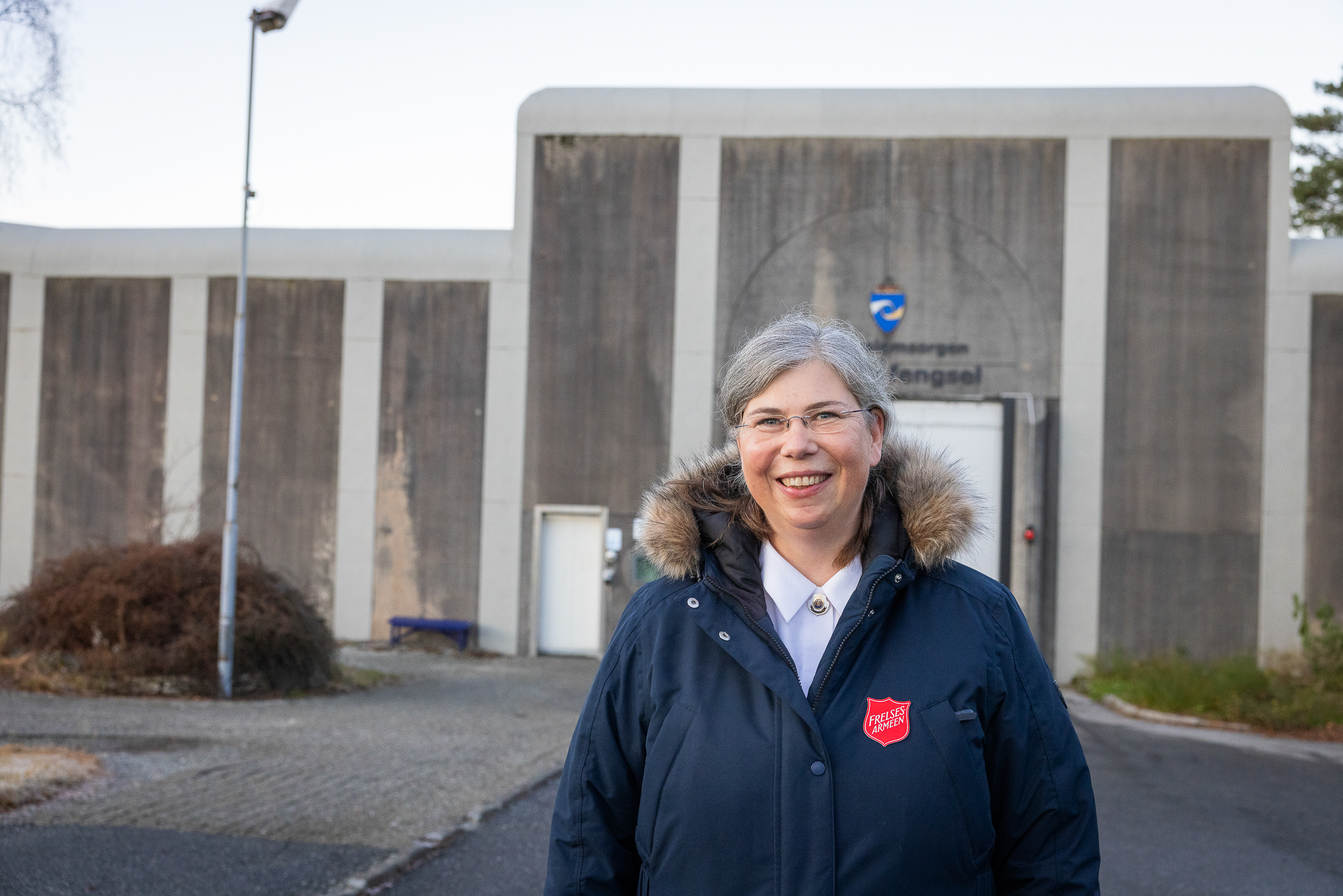 Kvinne i blå jakke med logo fra Frelsesarmeen står ute foran en febgselsmur.