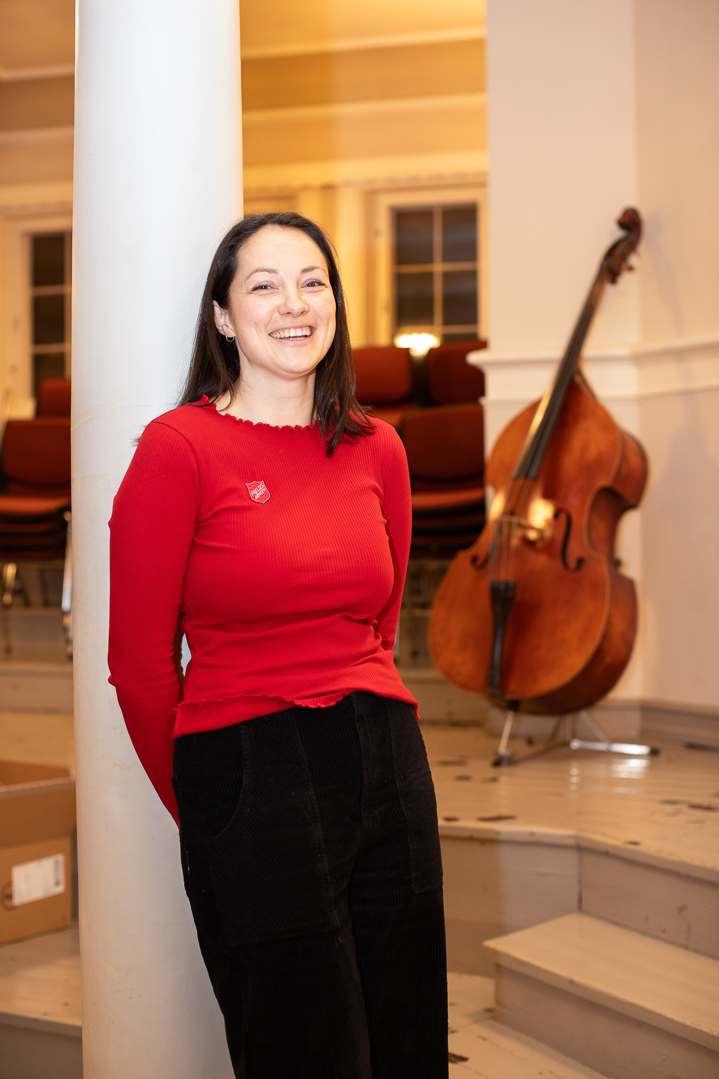 Kvinne i rød genser med Frelsesarmeens logo på brystet lener seg inntil en hvit søyle i betong. I rommet står en cello klar på et stativ opp noen få trappetrinn.