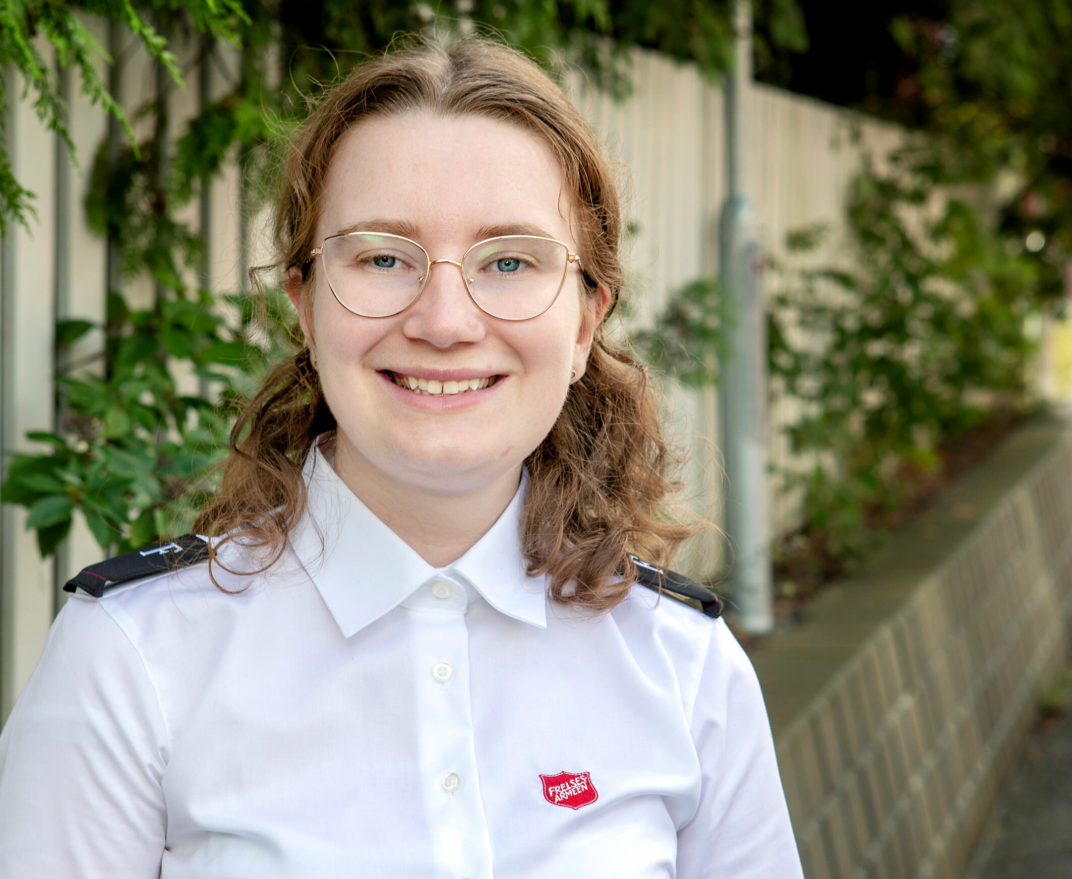 Foto: Ung kvinne i 20-årene med krøller og briller står i en hvit kortermet uniformsskjorte fra Frelsesarmeen. Hun står ute foran en mur og et gjerde med litt grønne vekster.