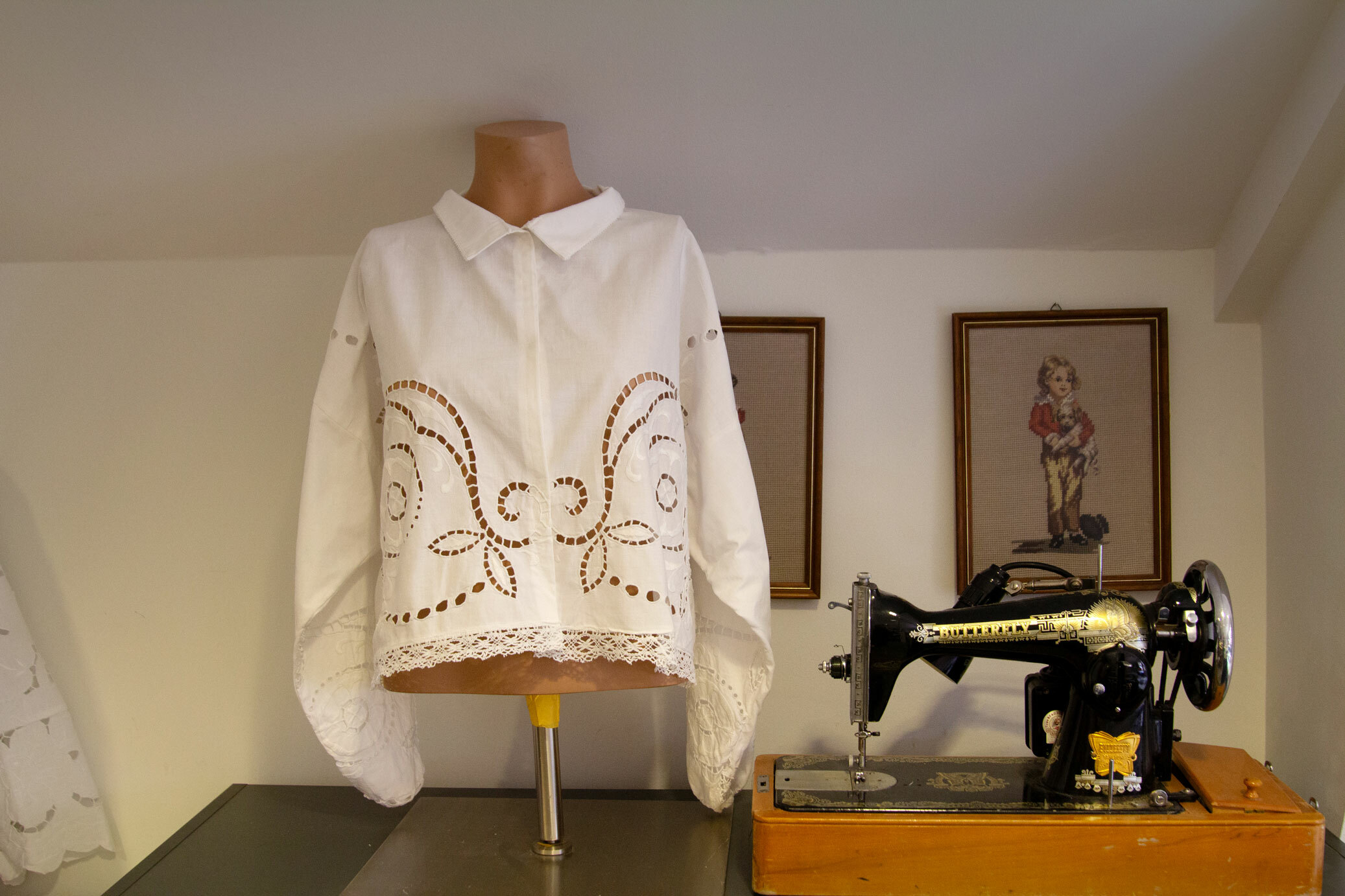 Foto: Hvit blondebluse står til utstilling på en byste. I bakgrunnen står det en eldgammel, håndsveivet symaskin og det henger et par broderte bilder.