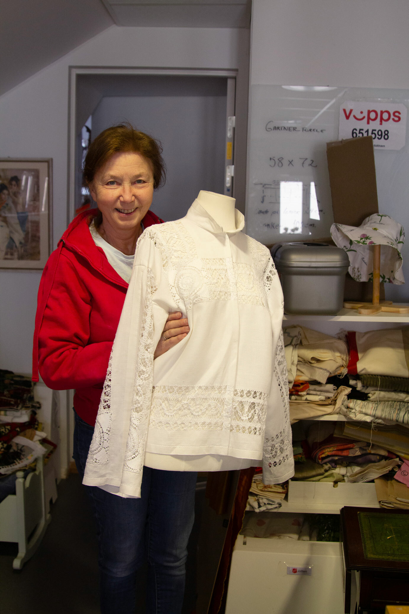 Foto: Kvinne sent i 50-årene med mørkt hår og rød jakke holder fram en utstillingsdukke (uten hode) som har på seg en vakker, hvit blondebluse.