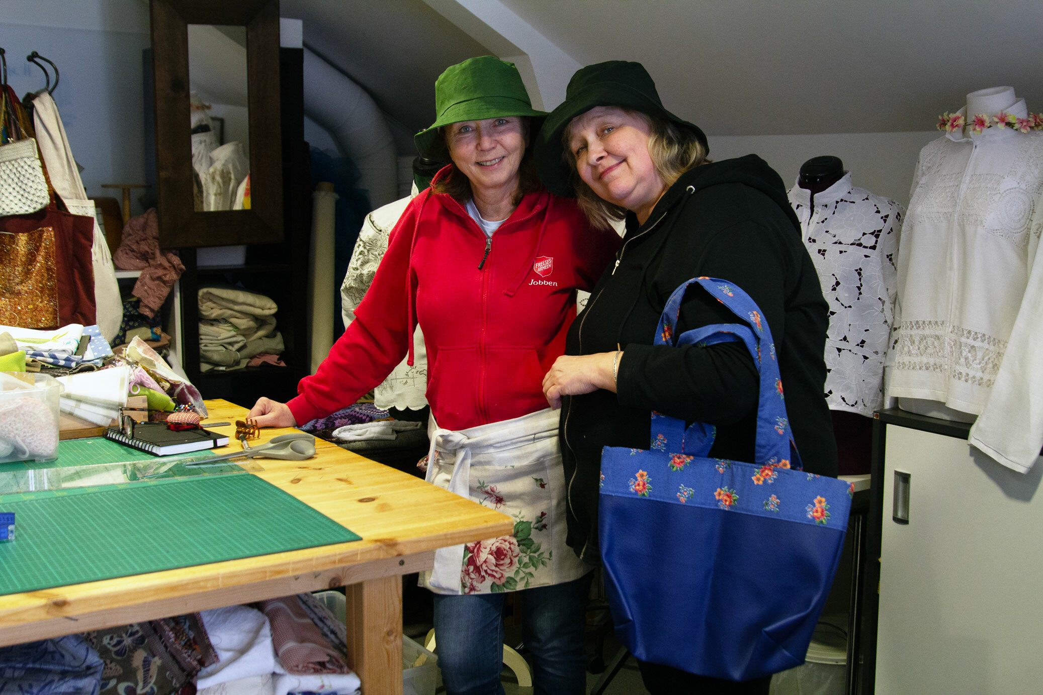 Foto: To kvinner i slutten av 50-årene med hver sin bøttehatt. Den ene er mørk i håret, den andre er blond. Den ene holder et nett og den andre har på seg et blomstrete forkle.