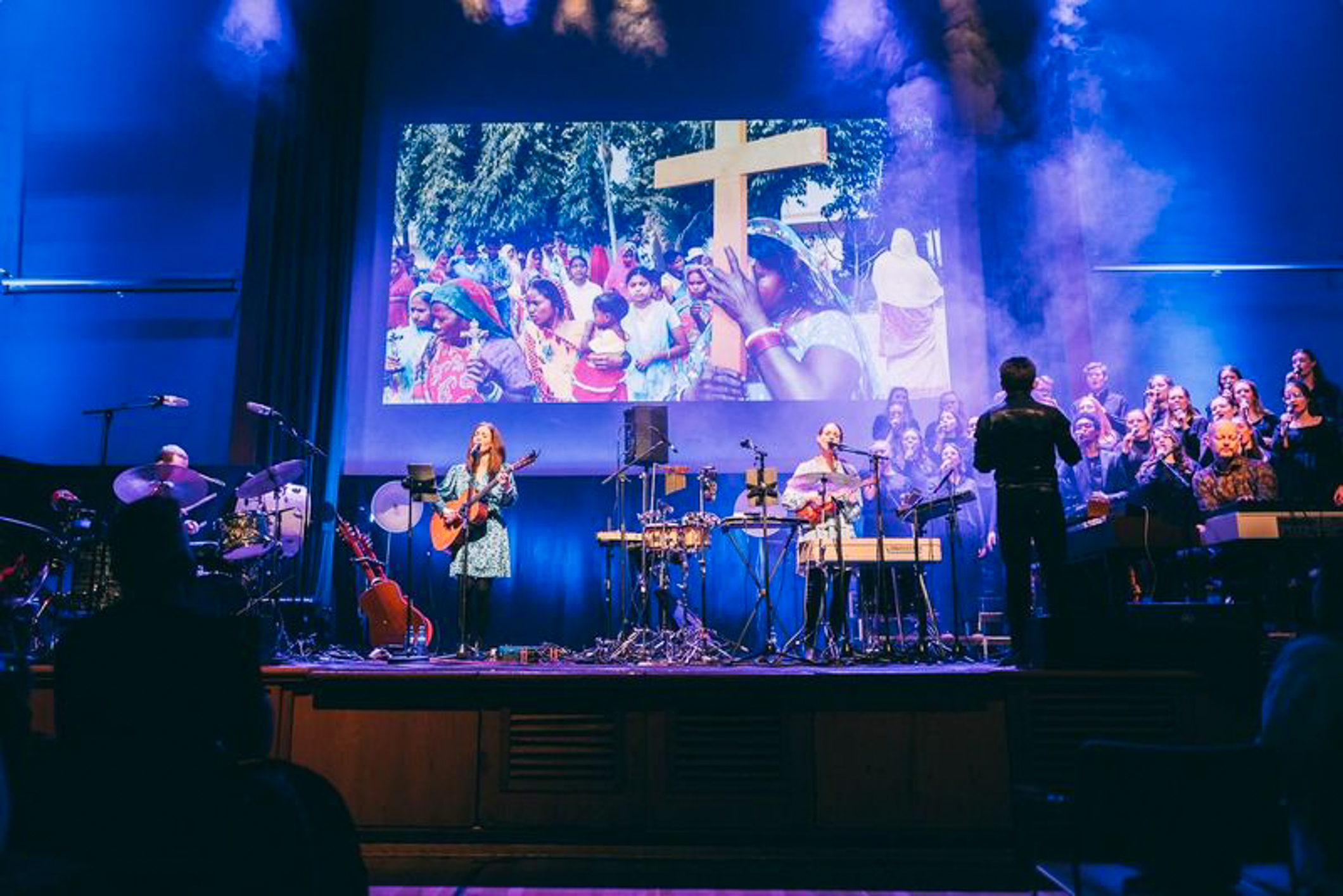 Bilde av en storskjerm med et bilde av mange mennesker i et fjernt land som bærer et stort kors. Foran på scenen står et par damer på hver sin gitar. Ute på høyre side står et kor og en dirigent.