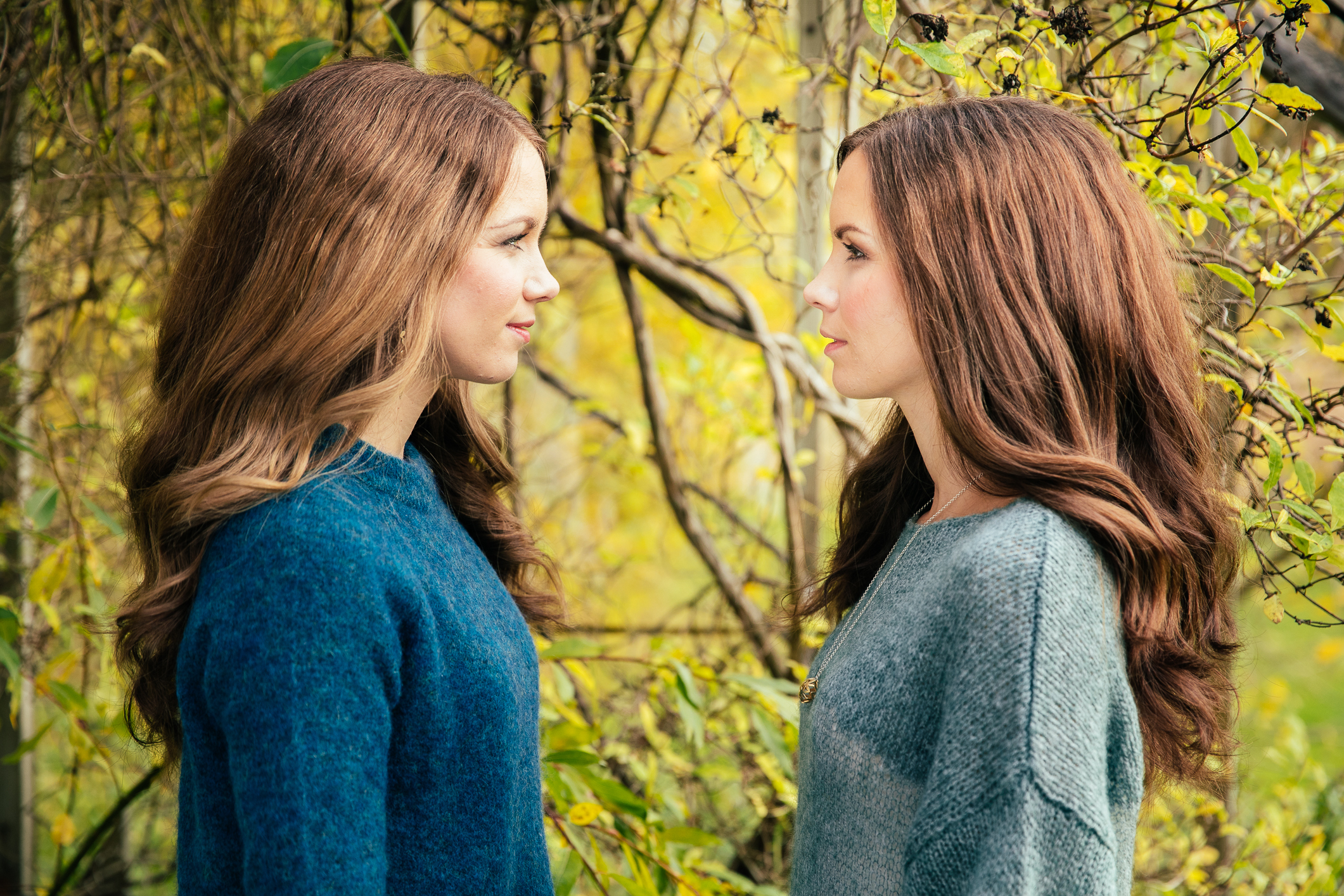 To prikk like søstre står ute i naturen med hver sin ullgenser på. De stirrer på hverandre og er tatt i profil.