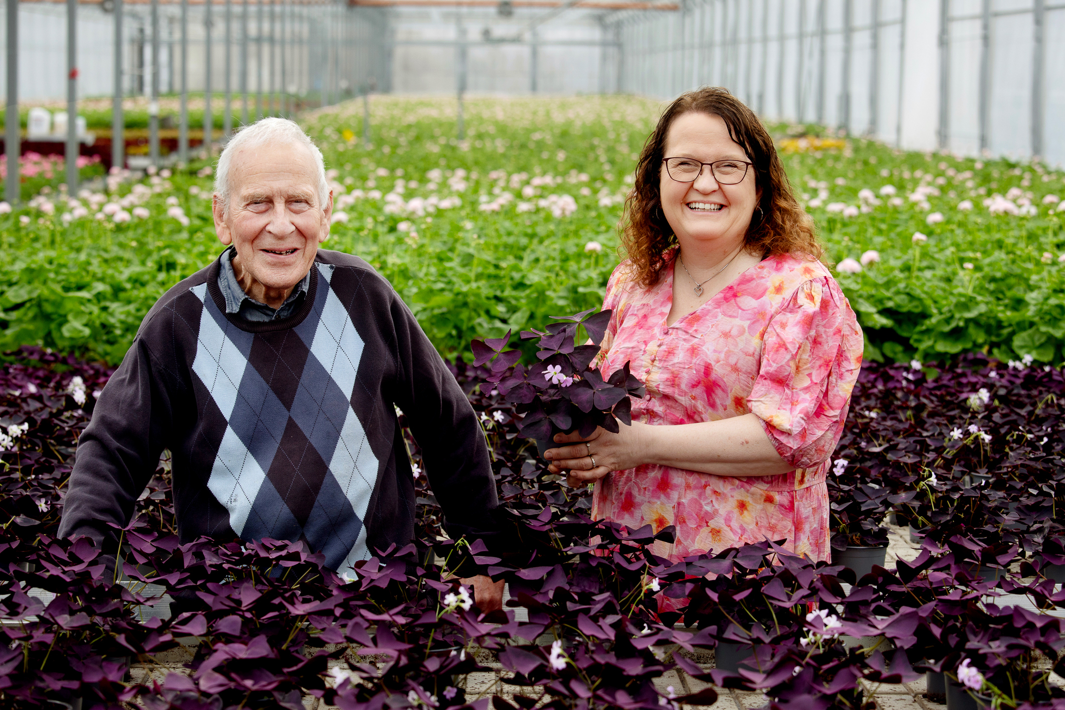 Eldre mann og yngre kvinne står i et blomsterhav inne i et drivhus.