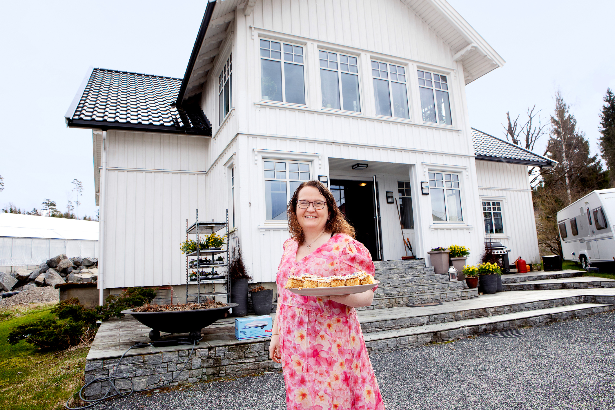 Kvinne i blomstrete kjole står med et kakefat foran en hvit sveitservilla.